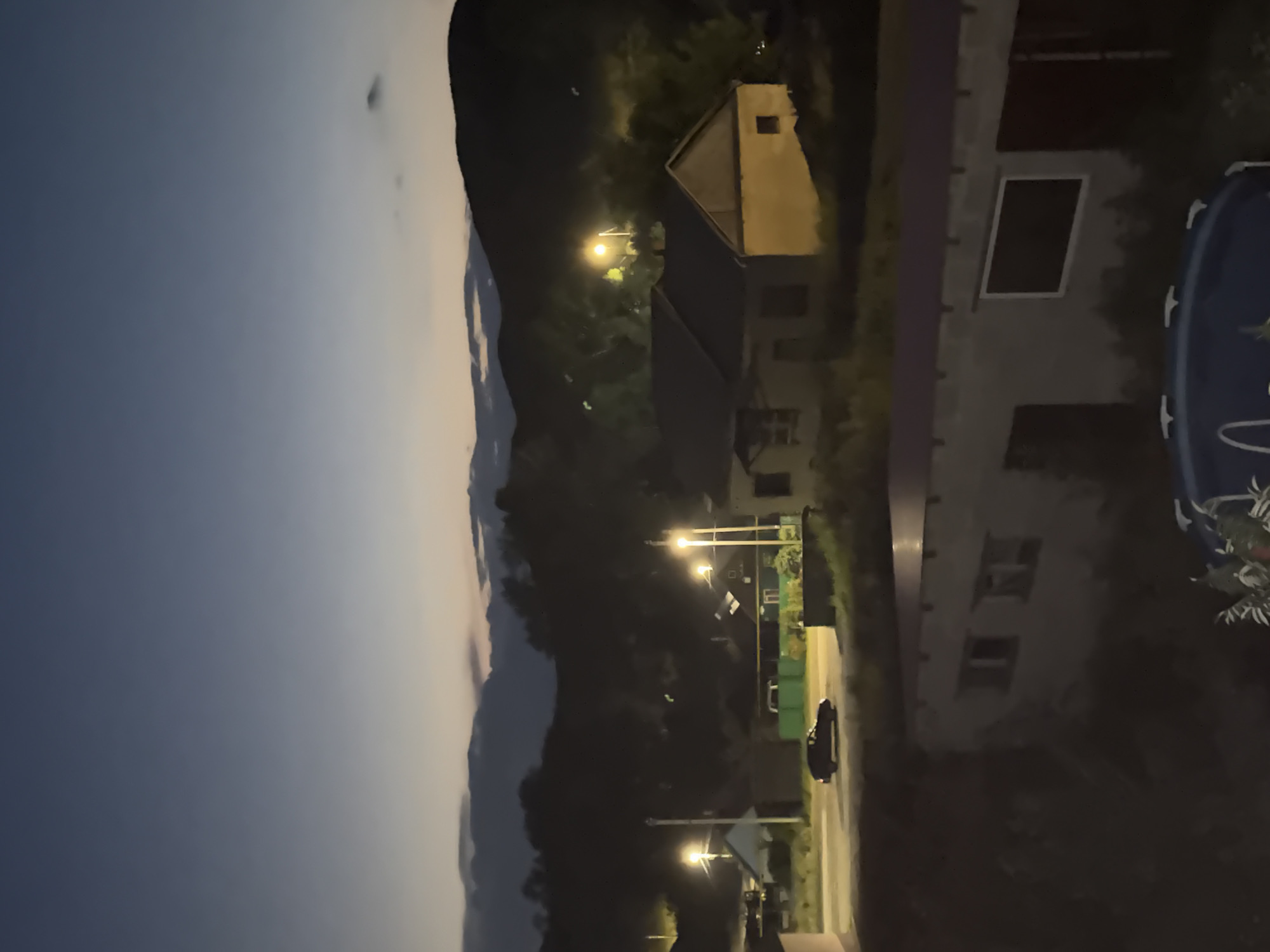 This image depicts a twilight scene in the city of Mednogorsk, Russia. The sky is a gradient of deep blue at the top, fading to a lighter, yellowish hue near the horizon, with faint clouds visible. In the middle ground, a rural village landscape is illuminated by streetlights. Several houses with pitched roofs are visible, some with lights on inside. A dark-colored car is driving on a paved road in front of one of the houses. Streetlights cast a warm glow on the surrounding area, illuminating trees and other vegetation. In the background, dark, silhouetted hills or mountains rise against the twilight sky. In the foreground, a part of a building with windows is visible, and further down, a blue inflatable pool is partially seen. The overall atmosphere is calm and serene, characteristic of a rural evening.
