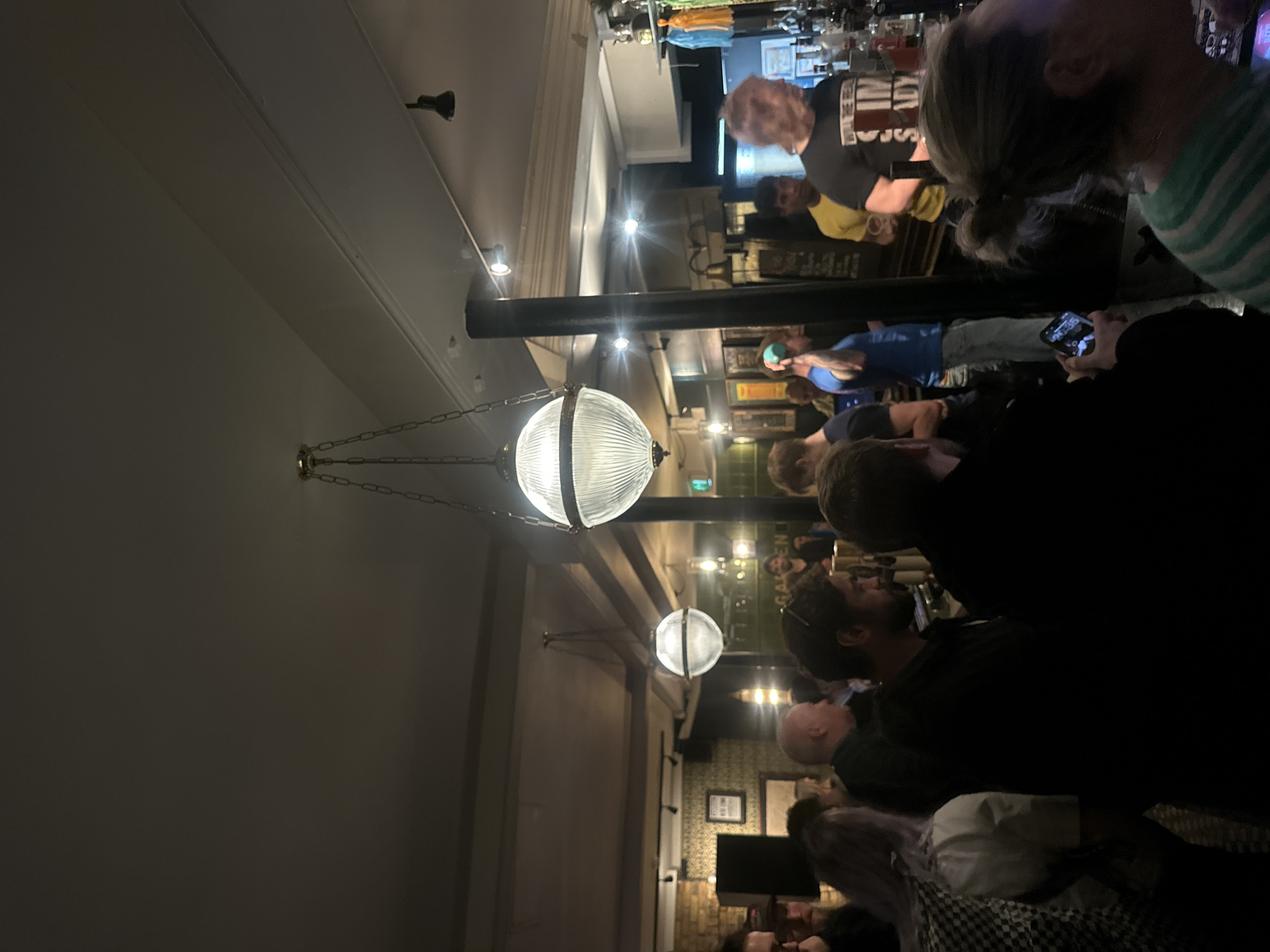 The image depicts the interior of a crowded pub. A row of people is gathered at the bar, engaging in conversation and looking at their mobile phones. A bartender is visible behind the bar, serving drinks. The pub is illuminated by hanging pendant lights and recessed ceiling lights. The pub's design incorporates dark columns and an assortment of framed pictures and advertisements on the wall behind the bar. The location appears to be a traditional London pub in the United Kingdom.