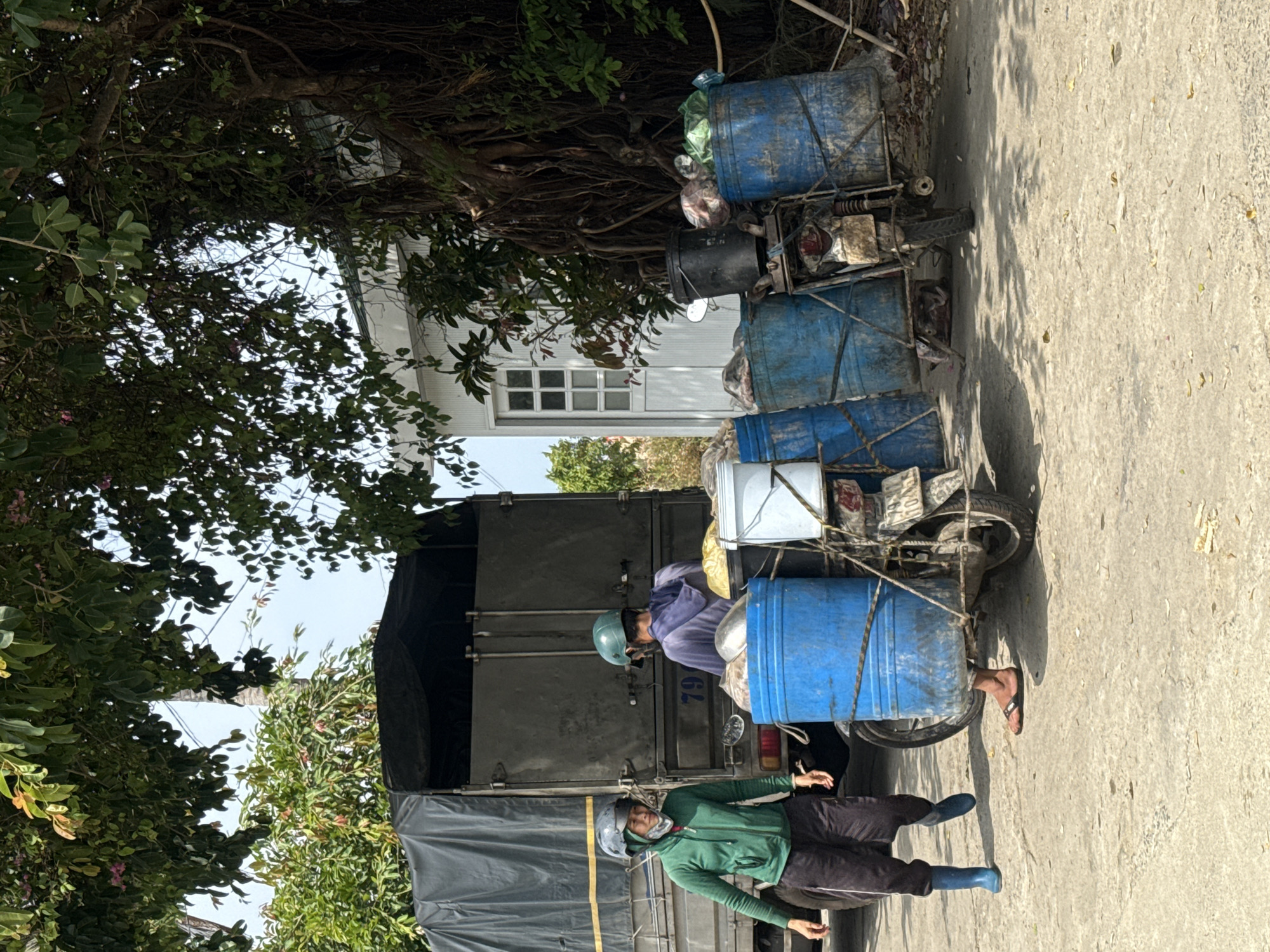 In the city of Bac Nha Trang Ward, Vietnam, an outdoor scene depicts two individuals near a large, dark-covered truck. To the right of the truck, a modified motorcycle is heavily laden with at least five large blue barrels, secured with ropes. One person, wearing a green jacket, black pants, and blue boots, stands to the left of the truck, facing towards the right. Another individual, in a purple hoodie and a green helmet, is positioned behind the motorcycle, appearing to interact with the barrels. The ground is a paved, textured surface, and lush green foliage from trees overhangs the scene, casting shadows. A white building with a window is visible in the background to the right. The overall impression is of a working environment, possibly involving transport or delivery.