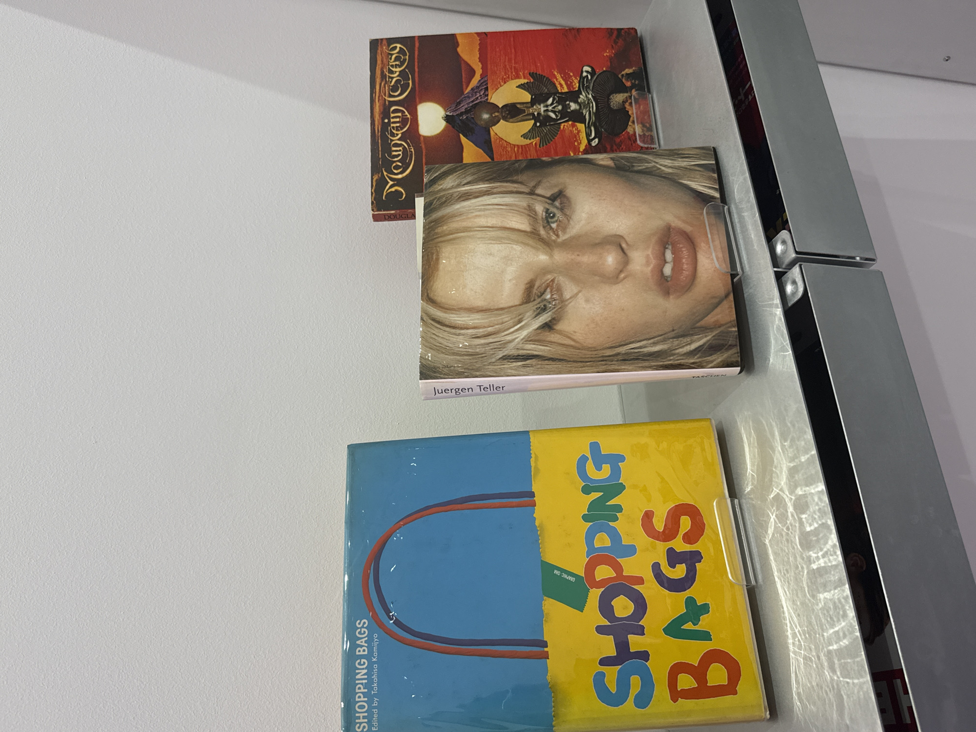 Three books are displayed upright on a polished metallic surface, likely a shelf or counter, against a plain white wall. The image is taken from a slightly elevated perspective, looking down at the arrangement.
The top book has a cover featuring the title "Mountain Ecstasy" in a decorative font. The artwork depicts a stylized figure resembling a totem or tribal deity against a desert landscape with a large sun or moon.
The middle book showcases a close-up portrait of a woman's face with light blonde hair, open lips, and a direct gaze. The name "Juergen Teller" is visible at the bottom of the cover, indicating the photographer or subject, along with the publisher "TASCHEN". This book is held in place by a clear plastic bookend.
The bottom book, also held by a clear plastic bookend, has a vibrant, colorful cover. The left side is blue with a simplified illustration of bag handles in red and orange. The right side is yellow with the bold, multicolored text "SHOPPING BAGS" arranged vertically. Below the main title, smaller text reads "Edited by Takahisa Kamijyo" and "GRAPHIC-SHA".
The metallic surface reflects light, creating dappled patterns. The setting appears to be an indoor environment, possibly a bookstore, gallery, or home display in London. There are no people visible, and the activity is simply the display of these art or photography books.