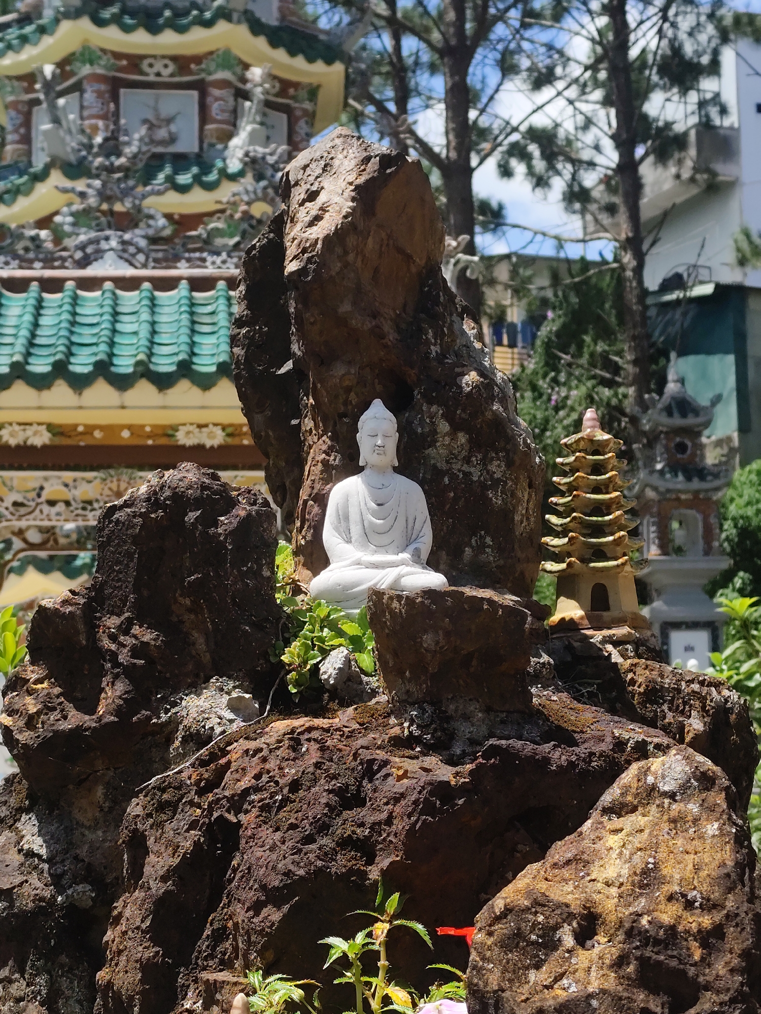 A white Buddha statue is sitting on a rocky outcrop with a miniature pagoda to its right, set against the backdrop of a building with green roofs and surrounding trees. The location is Da Lat, Vietnam, as suggested by the pagoda architecture. The Buddha statue is small, appearing to be made of white stone or plaster. It is in a meditative pose. The rocks surrounding it are large and dark, with some vegetation growing around the statue. The miniature pagoda is also made of stone or plaster, and it has multiple tiers. The background includes a pagoda-style building with a distinctive green tiled roof. Some trees and partial buildings are also visible. The scene is bathed in natural light, suggesting it is outdoors.