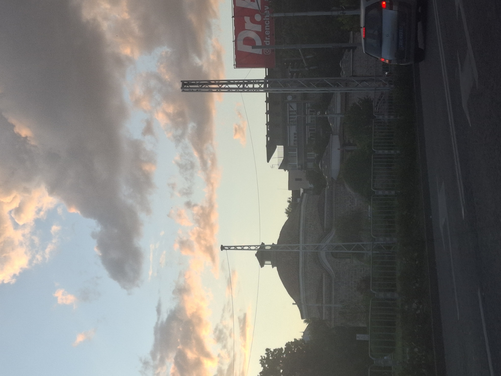 The image depicts an outdoor scene with a dramatic sky filled with clouds tinged with pink and orange, suggesting either sunrise or sunset. In the foreground, on the right side, a road is visible with white lane markings and a car with its brake lights illuminated. The car appears to be stationary or moving slowly. Adjacent to the road is a metal fence and some greenery.
In the mid-ground, there is a large, arched building with a brick facade and a domed roof. A metal structure, possibly part of a railway overhead line or a communication tower, stands in front of the building. Further back, there are several buildings, some with multiple stories and balconies. A prominent red billboard with white text is also visible, with the word "Dr." and what appears to be a social media handle "@dr.enchen". The overall impression is a somewhat urban or semi-urban environment during a colorful sky event. The orientation of the image is rotated 90 degrees counter-clockwise.