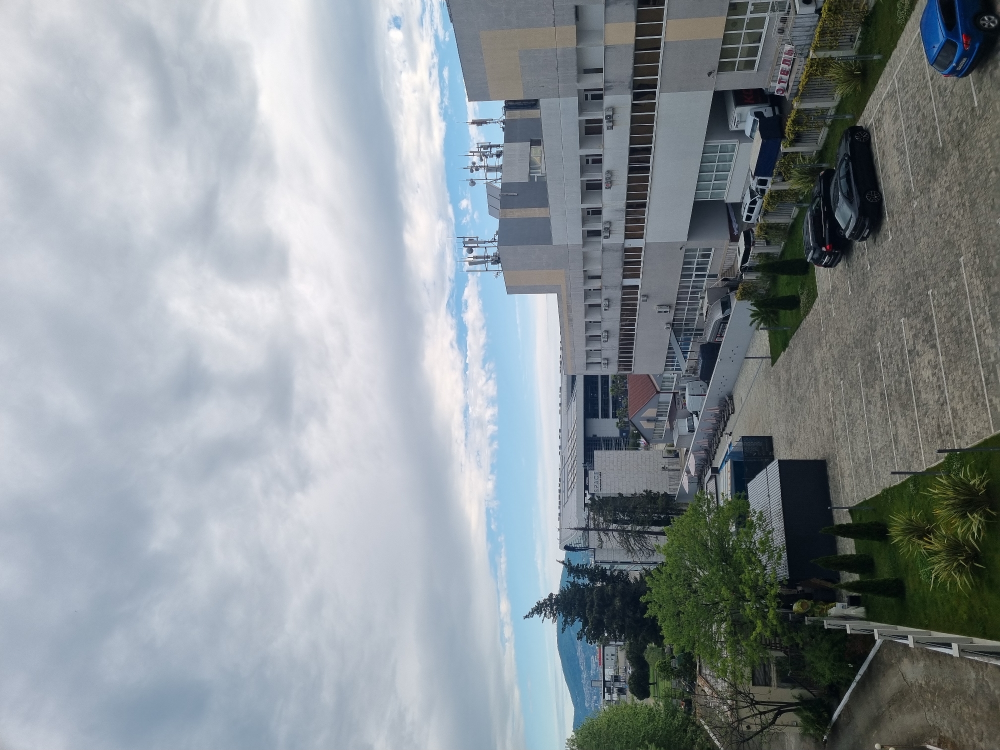The image is an aerial view of a parking lot and a multi-story building in Sochi, Russia. The parking lot has several cars parked, including a blue car and a few dark-colored vehicles. A building with multiple antennas on the roof is seen. A clear sky with clouds is in the backdrop, and a mountain is visible. Lush greenery surrounds the parking area. The side of the building has the word "OTEJD" written on it.