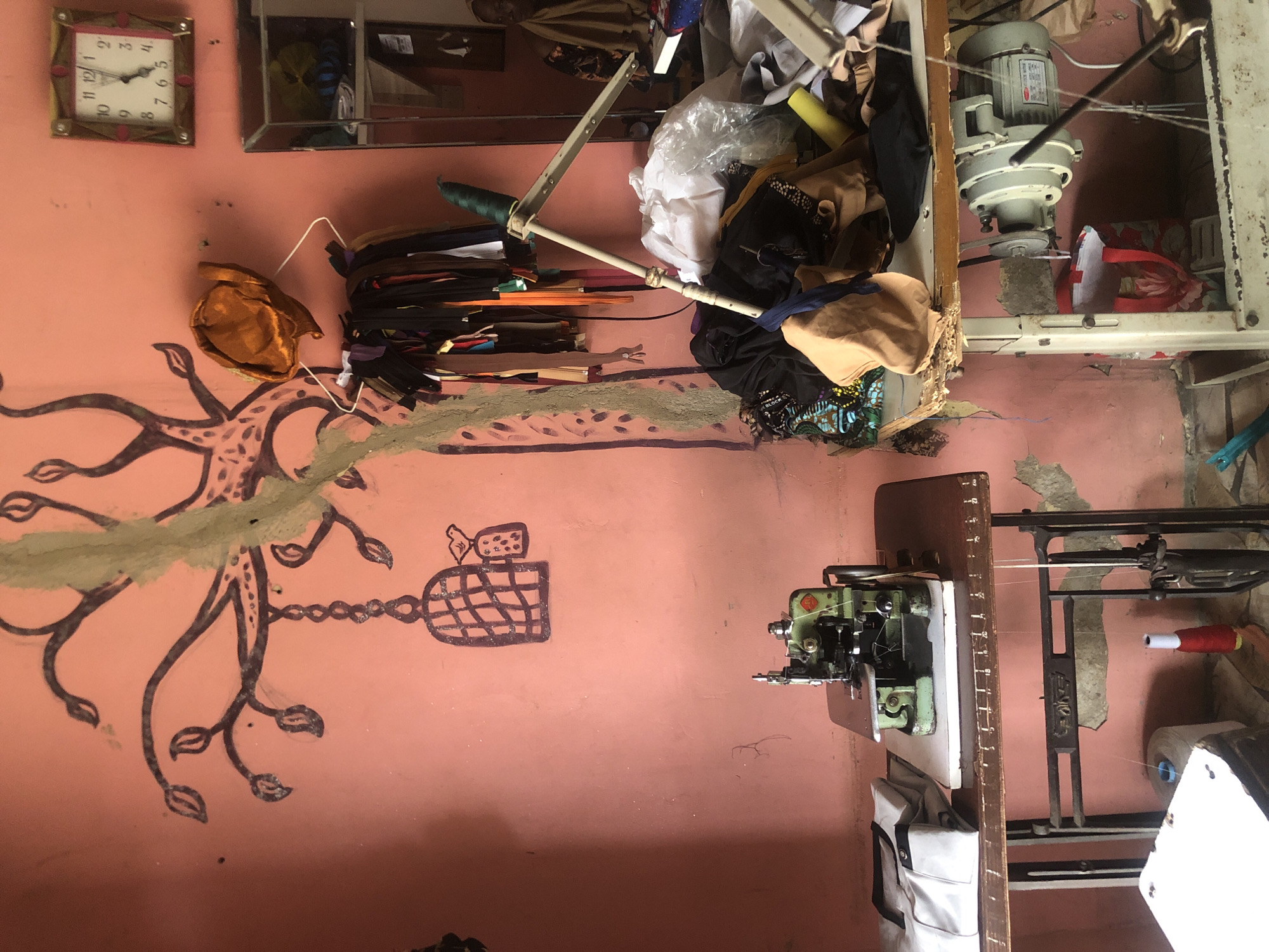 The image depicts a sewing workshop in Ado, Nigeria, characterized by a rustic and cluttered environment. A salmon-pink wall serves as the backdrop, adorned with a rudimentary black drawing of a tree with branches, leaves, a birdcage, and a small bird. To the right of the drawing, a square clock with a pink border displays the time as approximately 11:58. Below the clock, a mirrored cabinet is partially visible.
In the foreground, two sewing machines are present. The machine on the left is a green overlock machine situated on a dark wooden table with metal legs. Stacked on the table are folded white shirts with black trim. The machine on the right is a sewing machine with a motor mounted underneath a workbench cluttered with various fabrics, clothing items, and spools of thread. A large white plastic bag filled with fabric is also visible on this workbench.
Hanging from a metal hanger above the right workbench are numerous colorful zippers. A golden-brown fabric, possibly a hat or a head covering, is draped over the hanger. The floor appears to be concrete and shows signs of wear. Scattered on the floor are more fabrics, a red shopping bag, and a spool of red thread next to a white cylinder. The overall impression is of a functional but disorganized workspace.