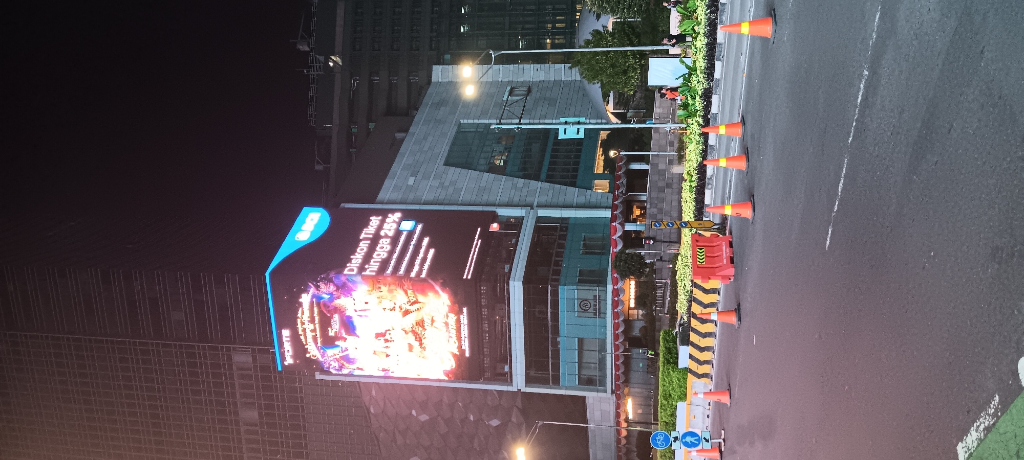 The image captures a nighttime street scene in the city of Central Jakarta, Indonesia, likely in a commercial district. Dominating the upper portion of the frame is a massive, illuminated digital billboard displaying a vibrant advertisement for "FORTE" and "BCI" with text in Indonesian promoting a "Diskon Tiket hingga 25%" (Ticket Discount up to 25%). Below the billboard is a multi-story building with modern architecture, featuring glass facades and horizontal window lines. The ground level of this building appears to have an entrance with red and white striped awnings.
In the foreground, a street is visible, marked with white lines. Orange traffic cones and a red construction barrier are placed along the roadside, suggesting some form of road work or traffic management. Pedestrian and bicycle lane signs are also present. Several streetlights illuminate the scene. In the background, other large buildings are visible, some with illuminated windows, indicating a dense urban environment. A few individuals can be seen in the distance near the buildings, but their activities are not discernible. The overall atmosphere is that of a busy urban area at night, with a strong emphasis on commercial advertising.