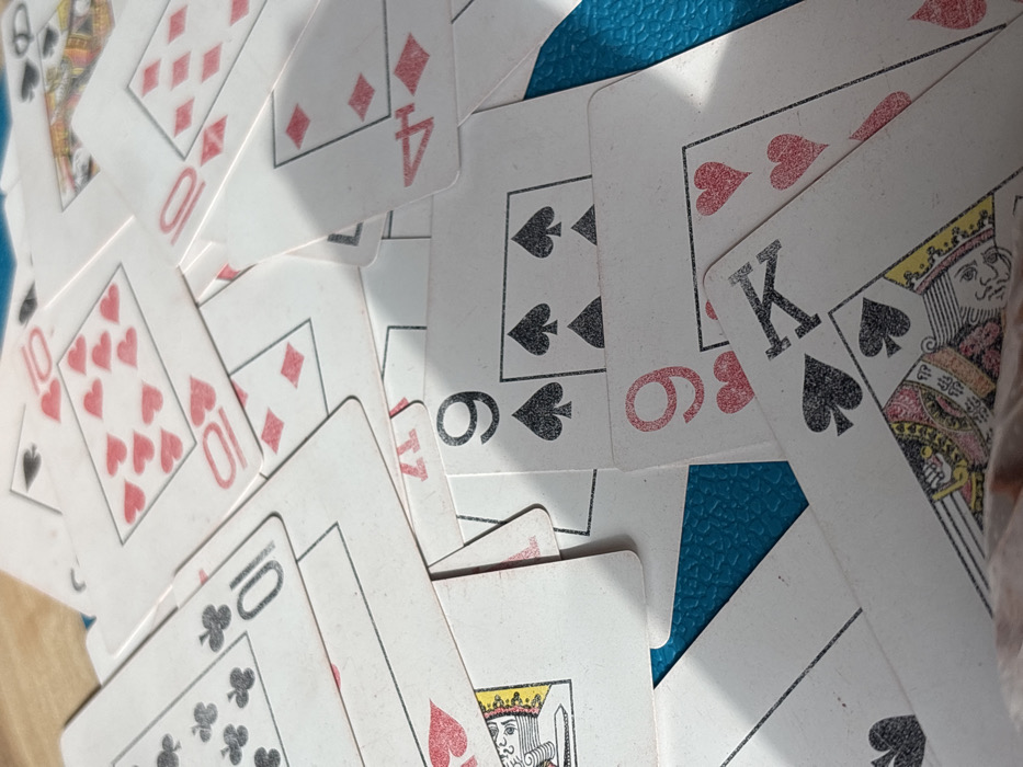 The scene is located in Ivanovka, Russia. The image provides a close-up, high-angle view of a pile of scattered and overlapping playing cards resting on a textured, teal-colored surface. The surface exhibits a consistent, fine-grain pattern, possibly a tabletop or mat. Illumination from the upper-left casts pronounced shadows across the cards, accentuating their contours and the layered arrangement. The cards are standard playing cards, primarily white-backed with red (hearts and diamonds) and black (spades and clubs) suits. Visible cards include a '10' of Clubs (upper left), a '10' of Hearts (upper center-left), a partially visible '10' of Diamonds (mid-left), a '4' of Diamonds (upper right), a '9' of Spades (mid-right), a '9' of Hearts (lower-center), a King of Spades (lower-left-center), and a partial Queen of Hearts (lower-left). The cards display minor wear and scuffing consistent with regular use. No individuals, actions, or interactions are depicted; the objects are static.
