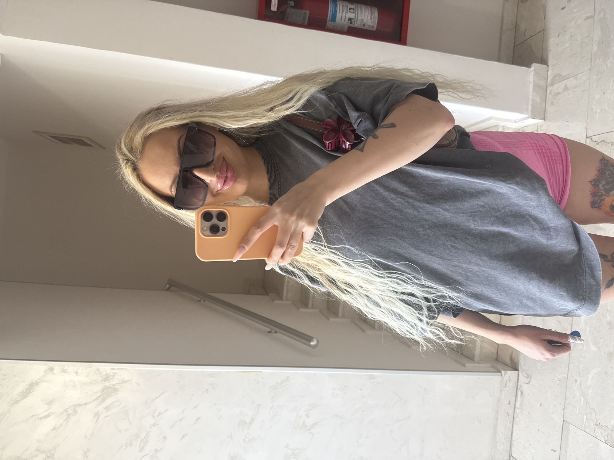 A blonde woman with long, wavy hair is taking a selfie in a hallway. She is wearing large, dark sunglasses, a loose grey t-shirt, and bright pink shorts. A small brown purse with a dark pink floral embellishment hangs from her shoulder. Her left hand holds a yellow smartphone, and her right arm is resting on her hip. She has tattoos visible on her thighs and forearm. In the background, there is a marble-patterned wall on the left, a staircase with a metal handrail, and a red fire extinguisher mounted on a wall to the right. The floor is tiled.