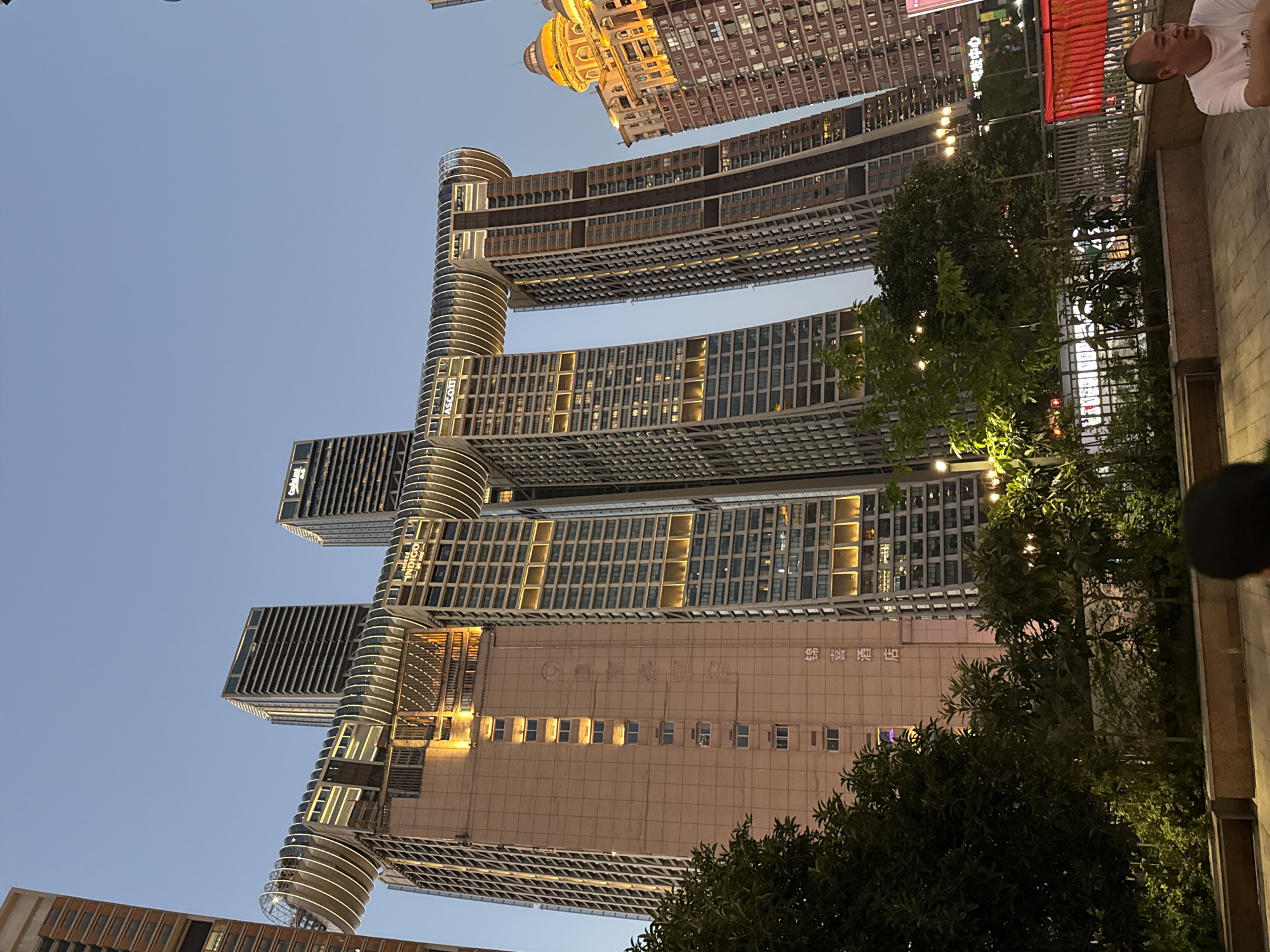 The image depicts the Raffles City Chongqing complex in Yuzhong District, China, viewed from ground level at dusk. The architectural marvel, designed by Safdie Architects, features four interconnected skyscrapers, with a horizontal "Crystal" structure spanning across two of them. The buildings are illuminated by warm, artificial lights, highlighting their modern design and intricate details. Green foliage and trees are visible in the foreground, creating a natural contrast with the towering urban structures. In the lower right corner of the image, a man wearing a white t-shirt is partially visible, looking towards the right. The sky is a clear, pale blue, typical of twilight.