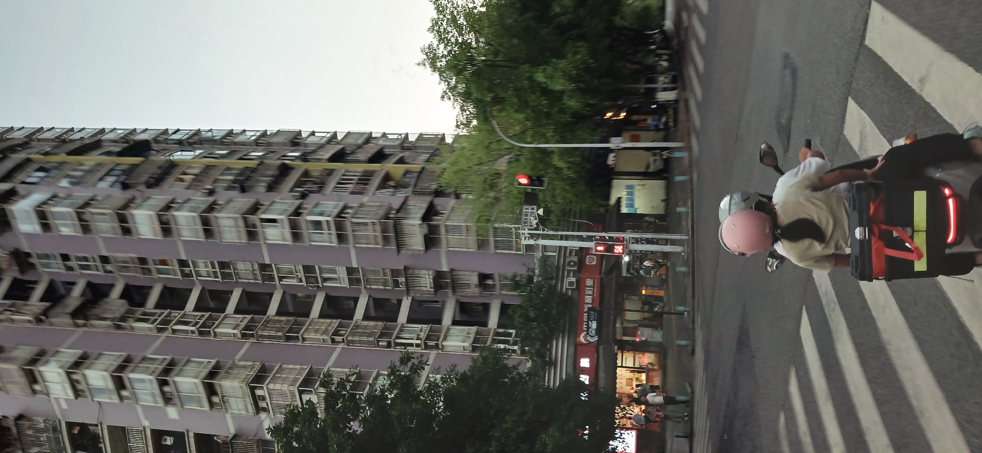 The image captures a street scene in Yuzhong District, China. A tall, multi-story building with numerous windows and air conditioning units occupies the left side of the frame. Lush green trees partially obscure the building and line the street. On the right side, there's a street with a crosswalk. A motorcycle carrying two individuals, both wearing helmets (one pink and one silver), is crossing the street. The motorcycle has a black storage box with red straps and reflective elements attached to the back. A traffic light and street signs are visible, along with storefronts and people on the sidewalk.