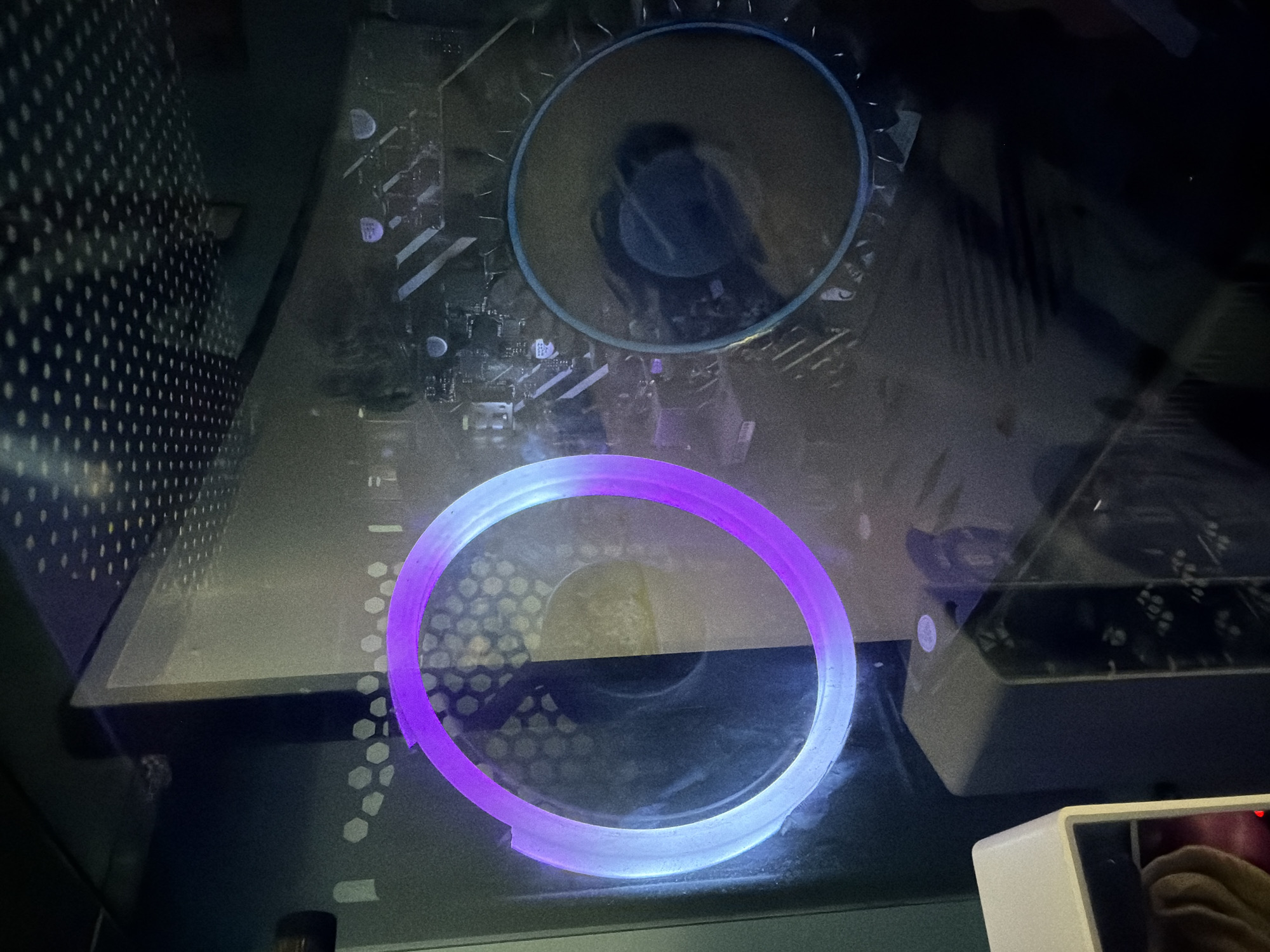 The image presents an interior view of an electronic enclosure, likely a computer case, observed through a transparent or translucent panel. The central focus is a vibrant, glowing circular light ring that exhibits a gradient from purple to light blue/white, illuminating the lower half of the frame. Above it, a larger, unlit circular element, possibly another fan or vent, is visible, with dark, indistinct internal components or reflections within its boundary.
The transparent viewing panel is noticeably dusty and shows various reflections, including faint patterns of electronic circuitry or a motherboard in the upper section. A hexagonal mesh pattern is visible below and to the left of the glowing ring, suggesting a fan grille or a textured surface. On the far left, a dark panel with a uniform pattern of small perforations indicates a ventilation area.
In the bottom right corner, a small white electronic device with a screen is partially visible, reflecting a hand, possibly that of the person taking the photograph, and a faint red light. The overall lighting is dim, accentuating the internal glow of the light ring. There is no discernible text visible in the image.