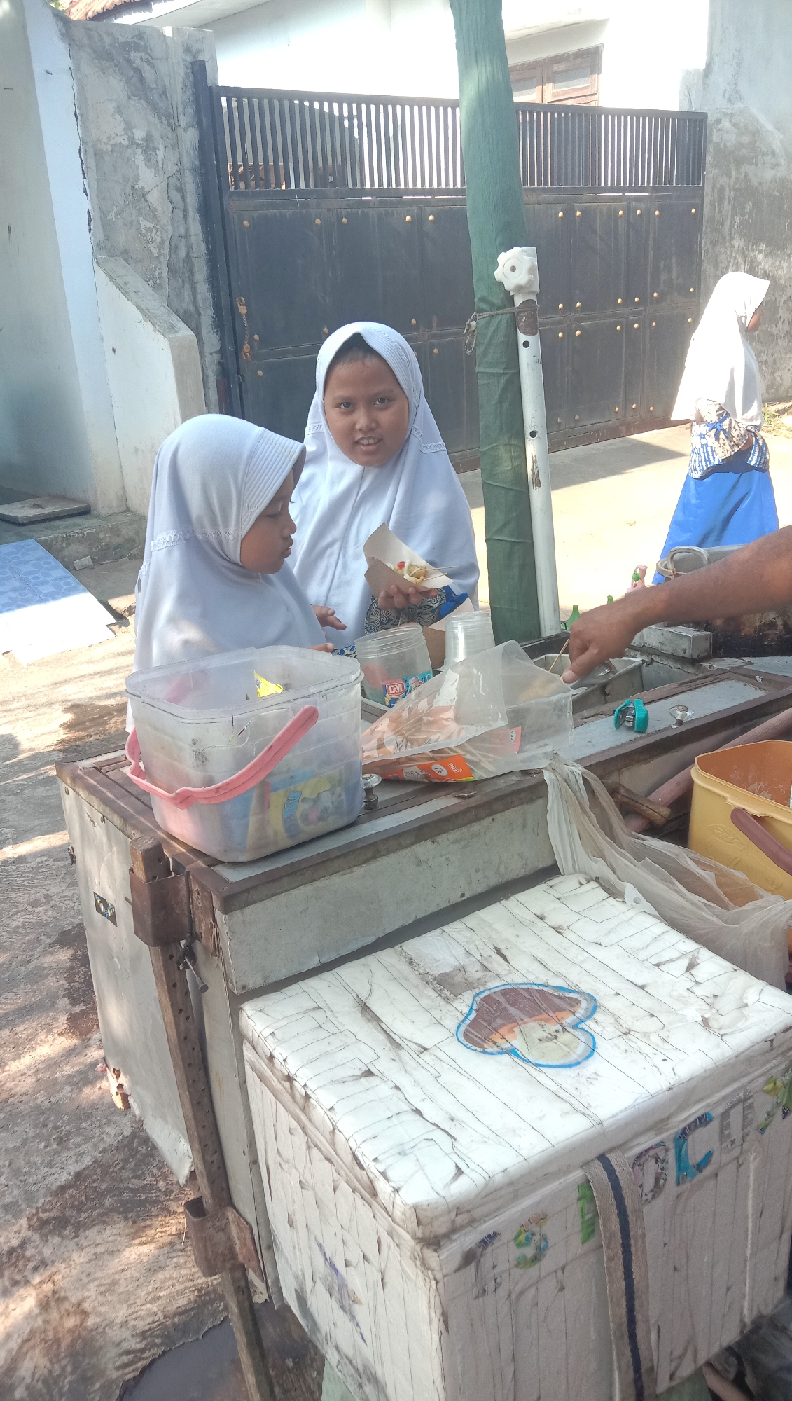 The image depicts two young girls in white hijabs standing near a street vendor's cart in what appears to be the city of Sidogiri, Indonesia. The girl on the right is holding a small paper cone filled with food, and she is looking towards the camera. The girl on the left is looking down and to the right, away from the camera.
The vendor's cart is old and appears to be made of metal and wood. It has a white, heavily textured lid on top, adorned with a faded cartoon sticker. Various containers and plastic bags are visible on the cart's surface, suggesting it is used for selling food or drinks. A person's arm, likely the vendor's, is reaching into the cart, holding a skewer or utensil.
In the background, a dark metal gate with vertical bars is visible, along with a section of a building and a tall, green-covered pole. Sunlight is casting shadows, indicating it is daytime. The overall environment suggests a casual, outdoor setting.