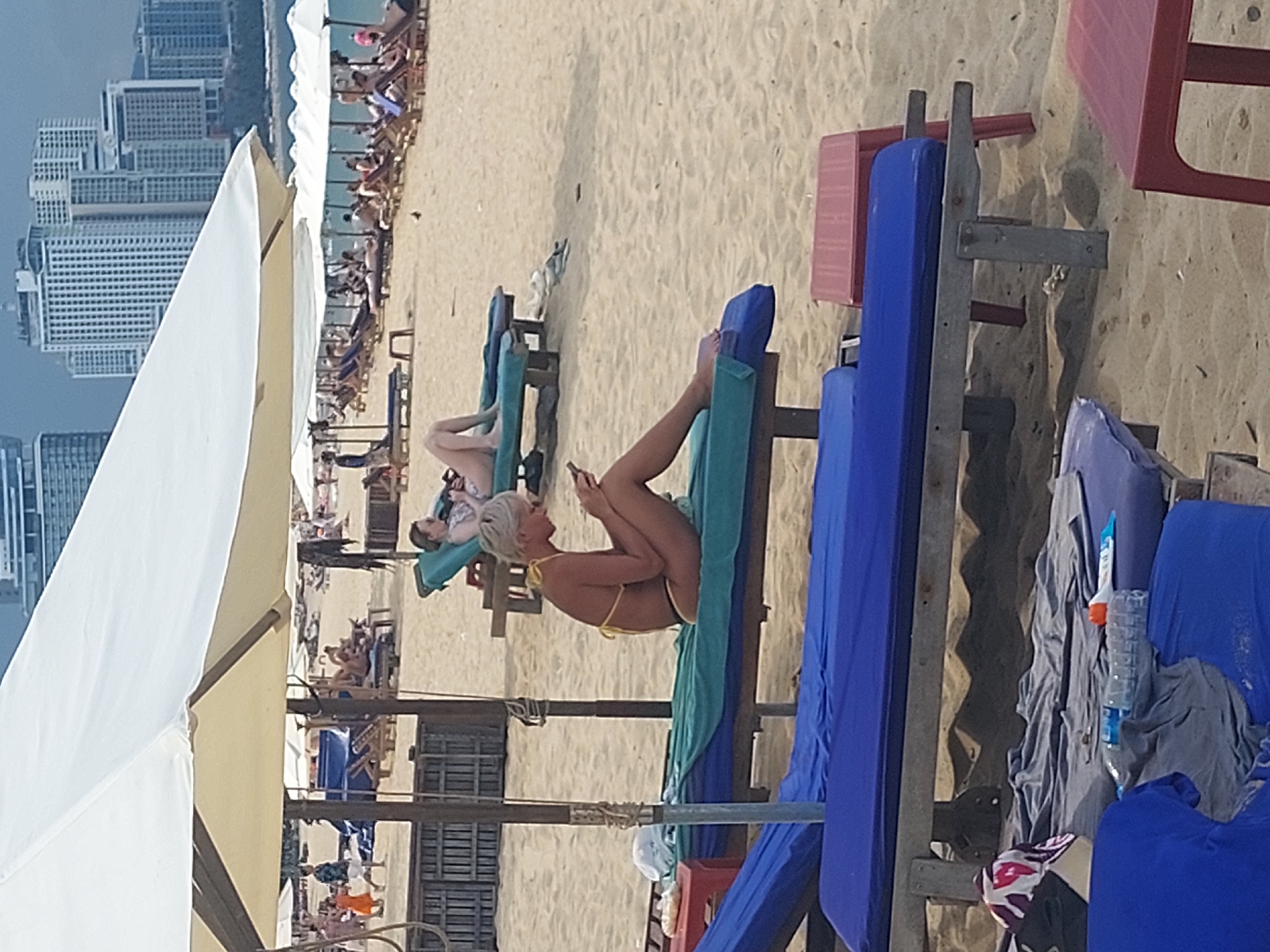 The image depicts a beach scene in Nha Trang, Vietnam. In the foreground, several blue and green lounge chairs are arranged on the sand, some covered with towels and a water bottle. A woman with blonde hair in a yellow bikini is seated on a green lounge chair, looking at her phone. In the middle ground, more lounge chairs and umbrellas line the sandy beach. In the background, tall buildings of Nha Trang are visible against a hazy sky. The overall scene suggests a relaxed atmosphere on a sunny day at the beach.