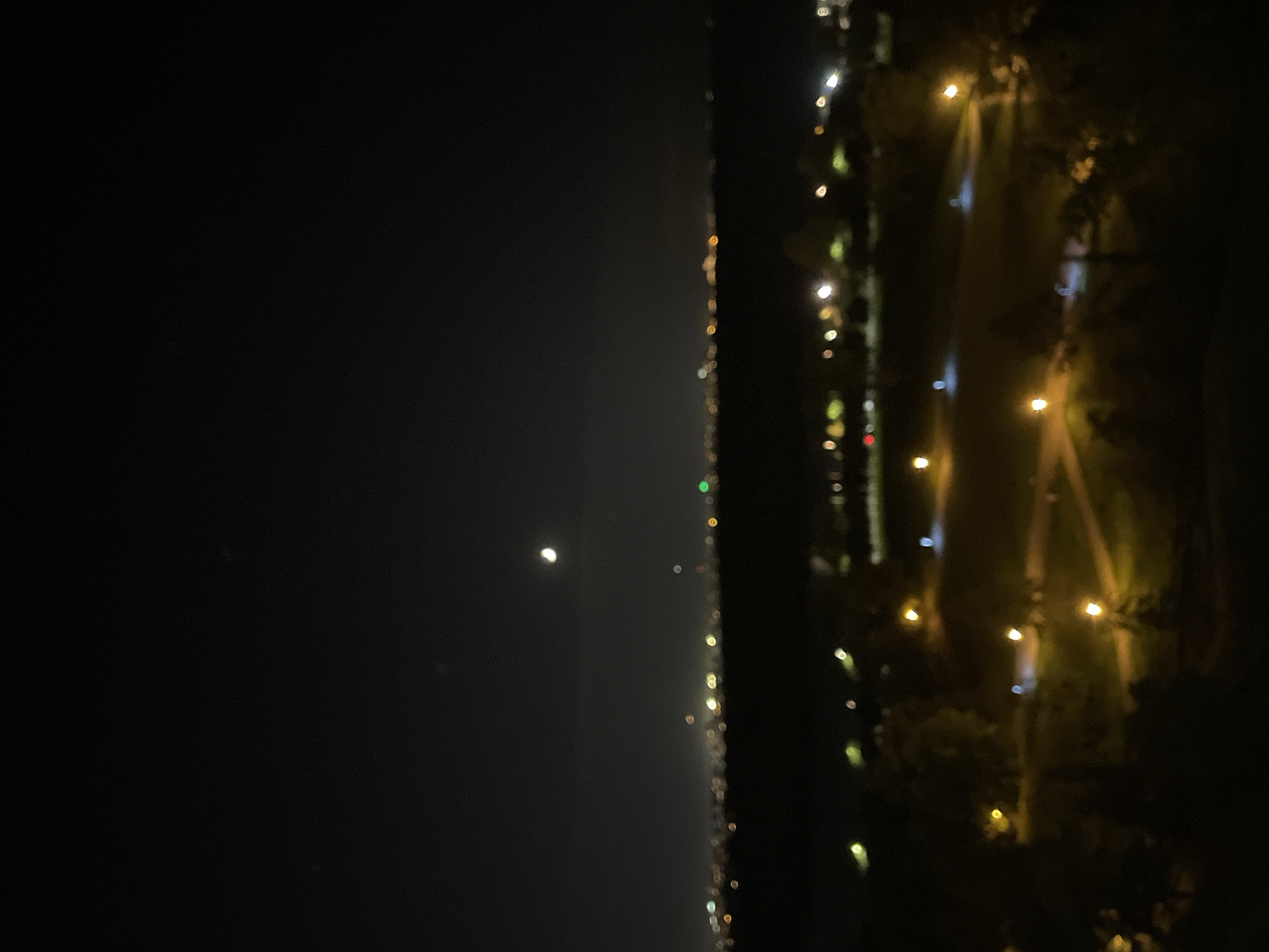 The image shows a night view of Belgrade, Serbia. On the left is a dark sky with a bright crescent moon. To the right of the sky is a line of lights, likely along the Sava river, representing the city skyline. On the far right are trees and lights that appear to be the illuminated paths and trees in a park or garden. The image is slightly blurred.