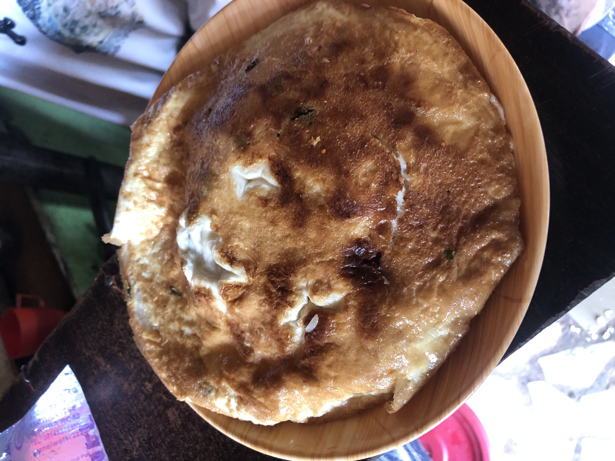 A close-up, overhead shot shows a golden-brown omelet on a light brown, circular plate. The omelet is irregularly shaped with a slightly raised, puffy center that has lighter, cream-colored sections, indicating undercooked egg. The edges of the omelet are darker and crisper. Scattered within the omelet are small green specks, possibly herbs, and tiny flecks of orange, possibly spices or dried chilies. The plate rests on a dark, textured surface, likely a wooden table. In the upper left corner, partially visible, is a clear plastic bag with purple text. Behind the plate and to the left, a red mug is visible. The background is out of focus, with hints of green and white fabric. There are no people or discernible actions taking place. The scene appears to be indoors, likely in a casual dining setting. The image does not contain any specific visual information to determine its location within the city of Kabong, Nigeria.