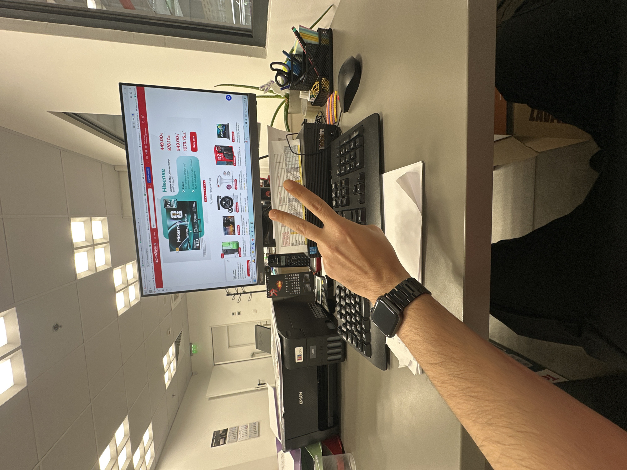 The image shows a workspace, likely in an office, in Lovech, Bulgaria. The workspace features a gray desk. In the center is a ThinkCentre keyboard and an Epson printer. A person's hand, wearing a black smartwatch, is raised in a peace sign over the keyboard. The computer monitor displays a Technopolis website page, advertising Hisense TVs. A pen holder with stationery items and a computer mouse are visible to the right of the monitor. The office is well-lit with fluorescent lights on the ceiling. A window is visible on the top left of the image, indicating the location is indoors.