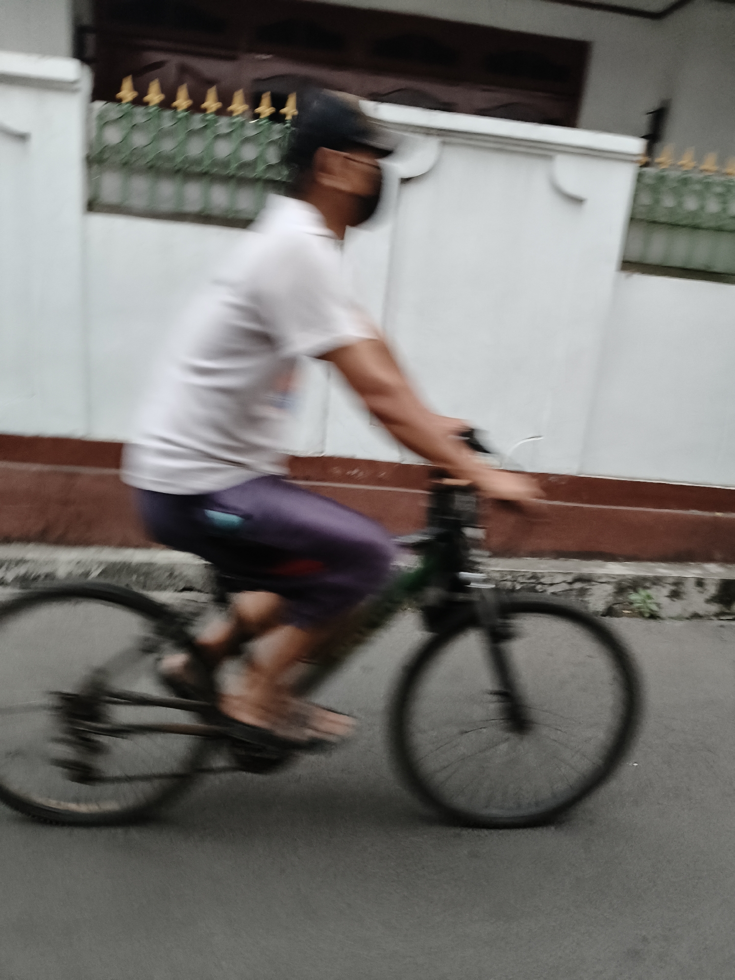 A person is riding a bicycle on a paved street in front of a building. The person is wearing a white shirt, purple pants, and a black cap, and a black mask. The building is white with a brown trim. The image is blurry due to motion. The location is South Tangerang, Indonesia.