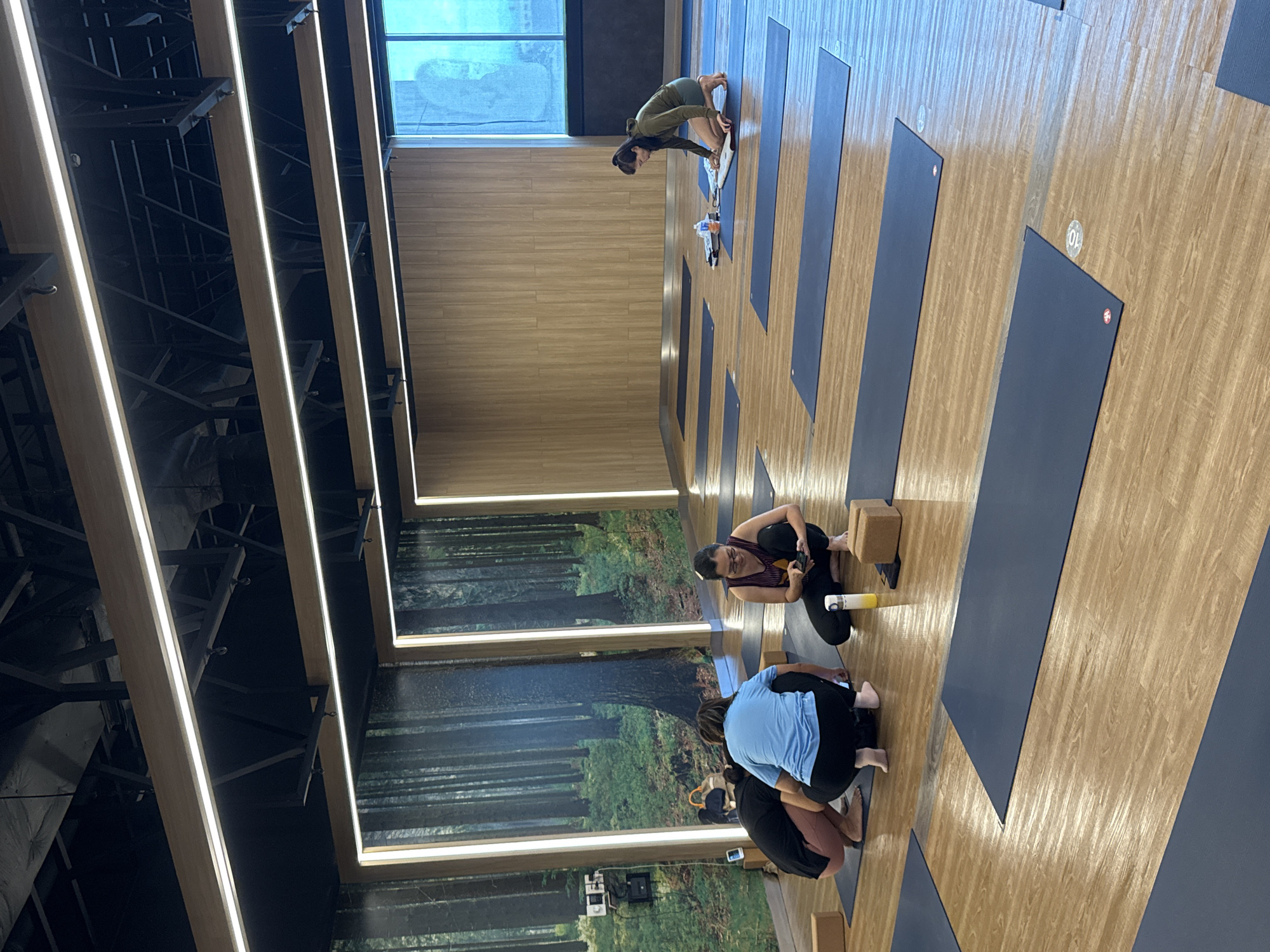 Four individuals are present in a yoga studio with a forest-themed mural on one wall and wood-paneled walls on others. The floor is wood-grained laminate with many dark blue yoga mats laid out. In the foreground, three people are crouched on mats. One person wears a black top and pink leggings, another a blue top and black leggings, and the third a purple tank top and black leggings. This third person is holding a cell phone. In the mid-ground, a person wearing a green long-sleeved shirt and shorts is kneeling on a mat, appearing to organize yoga accessories. The studio has modern lighting fixtures on the ceiling. There is a large window on the right side of the frame showing a bright sky, suggesting it is daytime. The image does not provide specific details about the city of Nong Pa Khrang, Thailand.
