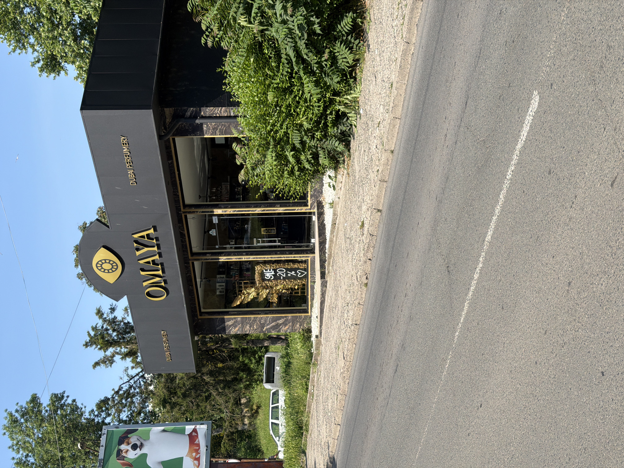 This daytime image, captured in Burgas, Bulgaria, presents an exterior view of the "OMAYA DUBAI PERFUMERY" storefront. The building features a dark, likely black or dark grey, facade with prominent gold lettering spelling "OMAYA" beneath a large gold stylized eye logo. Smaller gold text on the left and right sides of the main sign reads "DUBAI PERFUMERY." The storefront comprises large glass windows and doors, through which parts of the interior are visible. A white sign displaying "SALE -20% ❤️" is affixed to one of the windows, positioned in front of an elaborate golden decorative element resembling large wings or a statue.
In the immediate foreground, an asphalt road with a dashed white line runs parallel to a concrete curb. Beyond the curb, a paved sidewalk made of irregular, light-colored stones extends to the storefront. To the right of the perfumery, dense green bushes and foliage are present. To the left of the store, a large billboard displays an image of a smiling dog wearing sunglasses and a patterned skirt. Further in the background, a tree-covered hillside rises, and two vehicles—a white station wagon and a smaller grey or silver utility vehicle—are parked on a grassy incline. The sky is clear and blue. No individuals are visible within the frame.