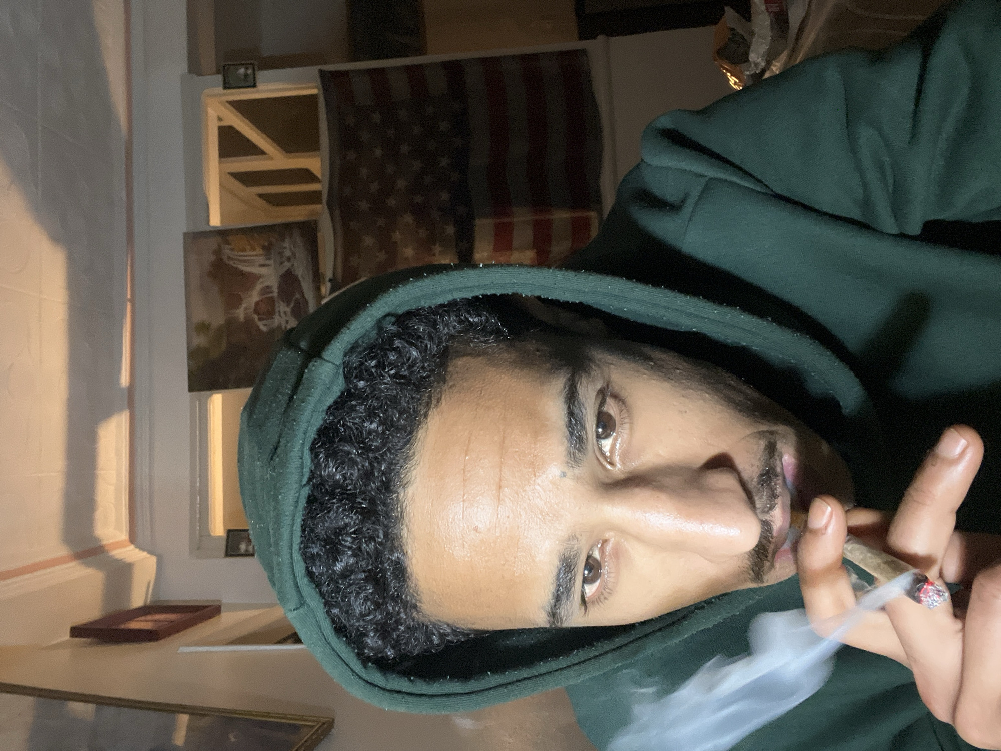 A man wearing a green hooded sweatshirt is holding a lit marijuana cigarette. Smoke is emanating from the tip of the cigarette. Behind him is an American flag and framed artwork hanging on a wall. The location is likely indoors, but the specific city and country details of Tanger, Morocco are not verifiable from the image.
