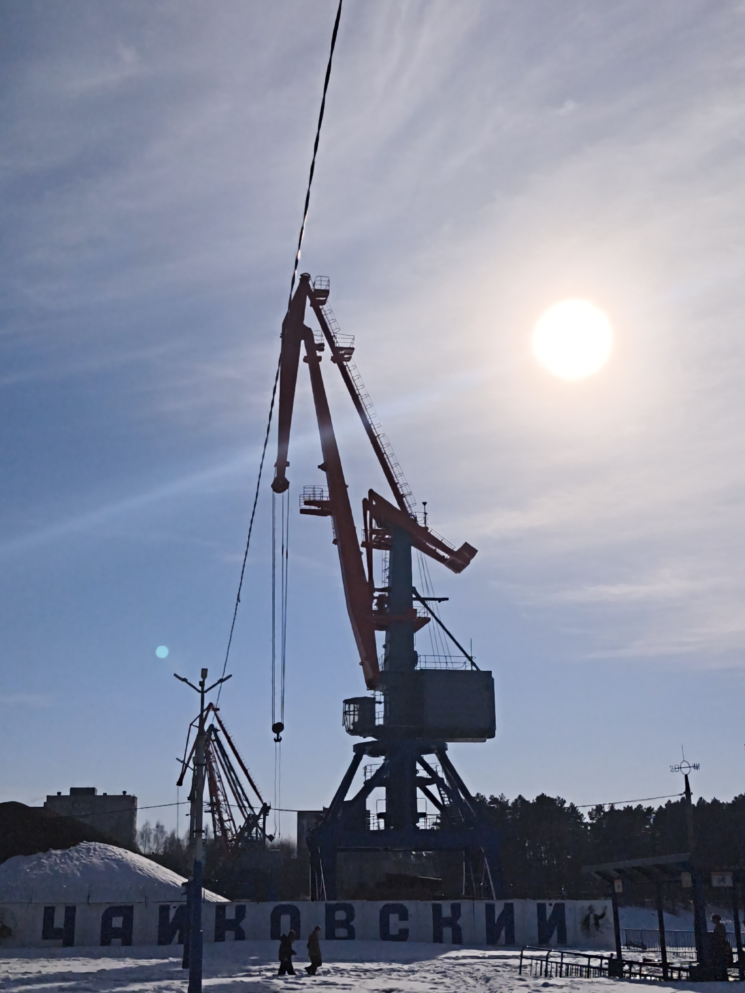 The image shows a sunny, winter day in what appears to be a port area in the city of Chaikovsky, Russia. Two large port cranes dominate the scene, silhouetted against the bright sky and sun. The cranes are of different sizes and are red and blue in color. Snow covers the ground. In the foreground, there is a sign reading "ЧАЙКОВСКИЙ," which is "Chaikovsky" in Russian. Two people are walking in the snow in front of the sign. In the background are trees and a building. A power line runs vertically across the sky on the left side of the image.
