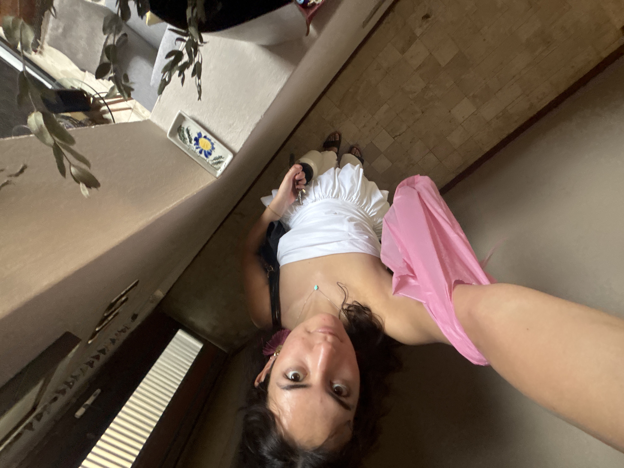A first-person perspective shot shows a woman standing in what appears to be a hallway or entryway. The woman is looking up at the camera, holding a set of keys in her left hand and a pink plastic bag in her right hand. She is wearing a white top with a ruffled skirt, brown sandals and a necklace with a light blue pendant.
The location is likely a building in Puebla, Mexico. The floor is tiled in a tan-colored rectangular pattern, the walls are beige, and decorative elements include a white ceramic piece with a blue floral design. A plant with green leaves is visible in the upper-left corner. In the lower-left corner, there is a dark brown paneled door or window with horizontal blinds.