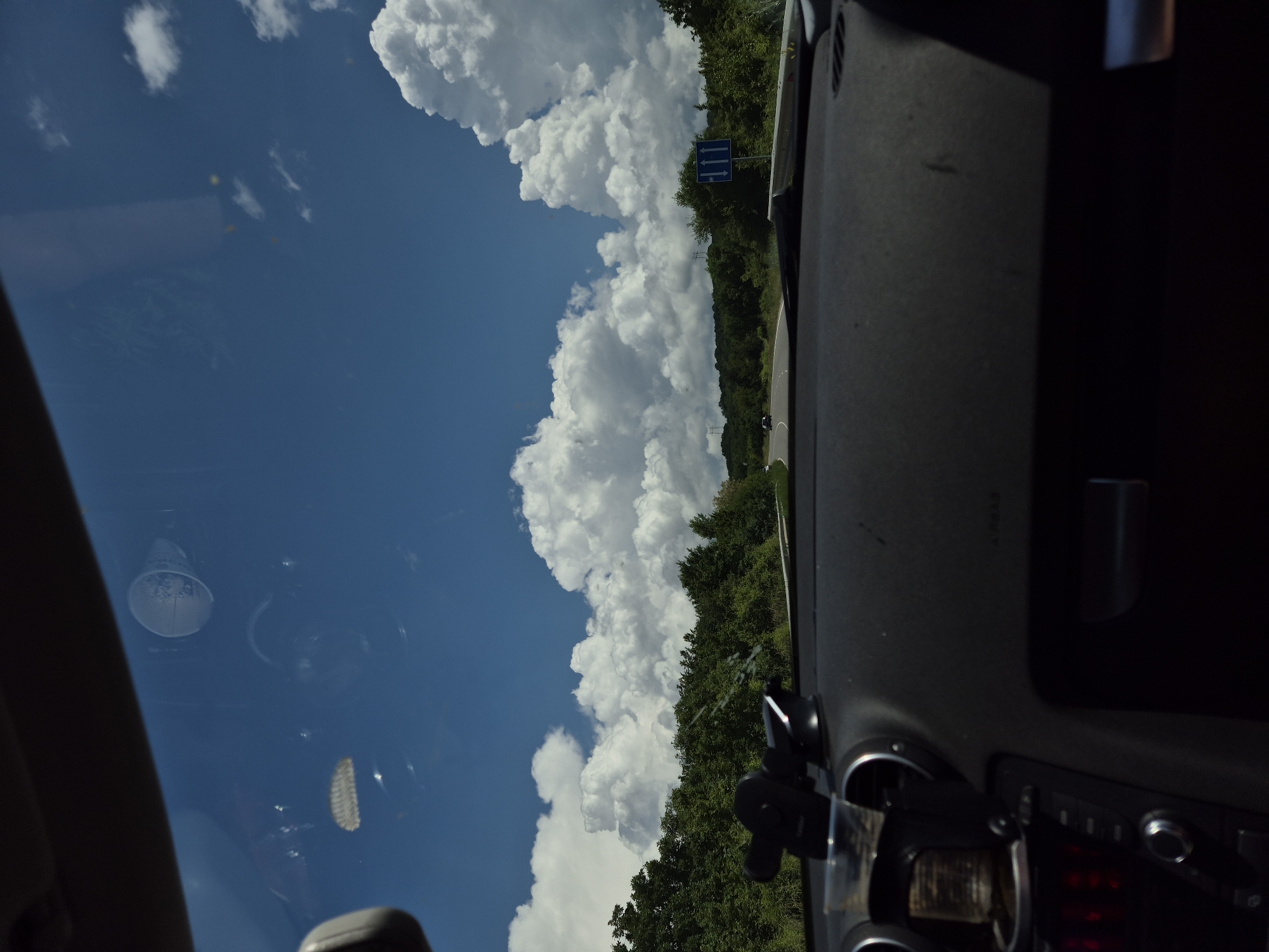 The image depicts a driver's perspective from within a vehicle, looking forward onto a two-lane asphalt road curving through a rural landscape under a bright blue sky with large, white cumulus clouds. Dense green deciduous trees and bushes line both sides of the road, extending into the distance. On the right side of the road, a blue rectangular road sign with three upward-pointing white arrows indicates traffic lane information. Faintly visible in the distant left are structures resembling electricity transmission towers above the tree line. The vehicle's dark-colored dashboard occupies the lower portion of the frame, showing a black suction-cup phone mount in the center-left, circular air vents, and part of a control panel with red illuminated elements on the lower left. Reflections on the windshield include a white disposable cup in the upper right quadrant and other indistinct shapes. The word "AIRBAG" is faintly visible on the passenger side of the dashboard. This scene is situated in Bogyovtsi, Bulgaria.