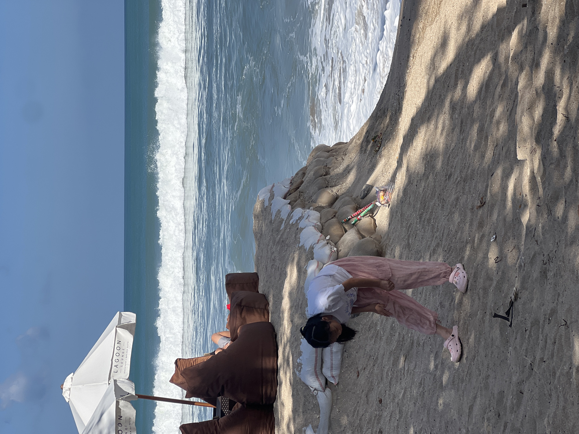 A young girl wearing a white t-shirt and pink patterned pants and pink sandals is bending over on a sandy beach in Kuta, Indonesia. She is looking down at something on the sand. Behind her, large brown beanbag chairs and white umbrellas from a beachfront lounge are visible. The ocean with white waves is in the background, and sandbags are stacked along the edge of the beach. The sky is clear and blue.
