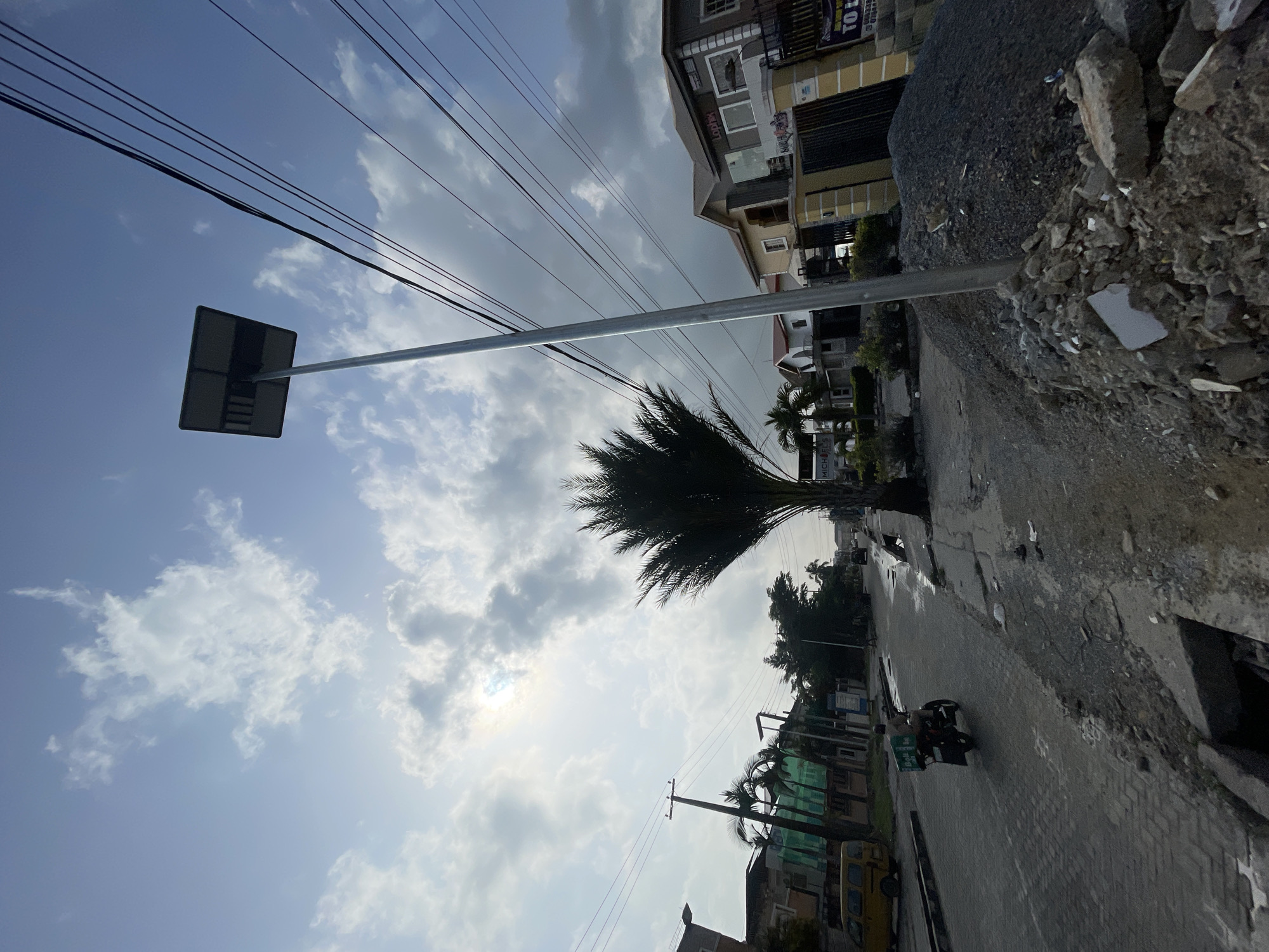 The scene captures a street in Moba, Nigeria. The road is paved with bricks and lined with buildings of various styles, including multi-story residential structures. Construction debris is piled on the right side of the frame, bordering the road. A palm tree stands prominently near the roadside. Electrical wires span overhead. A motorbike with a rider is visible on the street. A street sign is positioned on the left, reaching across the road. The sky is partly cloudy, with glimpses of sunlight.
