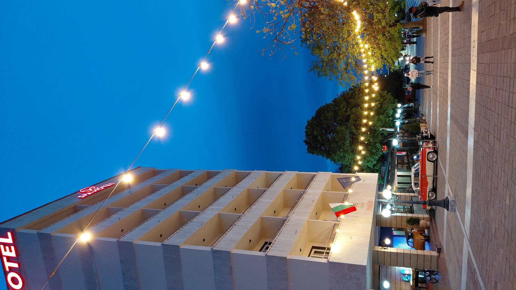 The image depicts an exterior view of the SPA Hotel Spartak in Sandanski, Bulgaria, at dusk. The hotel is a multi-story building with a modern design featuring a grid of recessed balconies. Red neon signage spelling "HOTEL" and "SPA HOTEL" is visible on the facade. A Bulgarian flag and another flag are displayed near the entrance. Strings of decorative lights are strung above the street, creating a festive atmosphere.
On the paved street in front of the hotel, there is a vintage two-tone red and white car parked. Pedestrians are walking on the street in the background. The sky is a deep blue, indicating the evening hour. Trees are visible along the street, some illuminated by the string lights. Street lamps provide additional lighting.