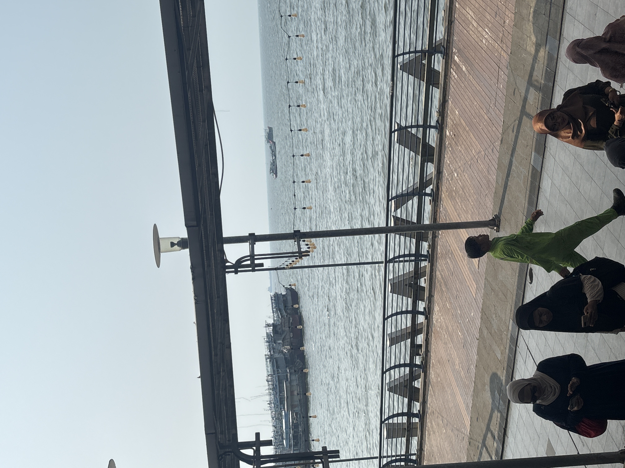 The image depicts a scene near the Sunda Kelapa Harbor in North Jakarta, Indonesia. In the foreground, several individuals are visible on a paved walkway. Two women wearing hijabs are looking at their phones, and another person, possibly a child, is walking in a bright green outfit. Further back, more people in hijabs are present, one of them seated. The walkway overlooks the water, where numerous boats and ships are docked or in transit. A decorative metal railing separates the walkway from the water. String lights are suspended over the water, and the sky is a hazy blue. The overall atmosphere suggests a public area near a busy port on a bright day.