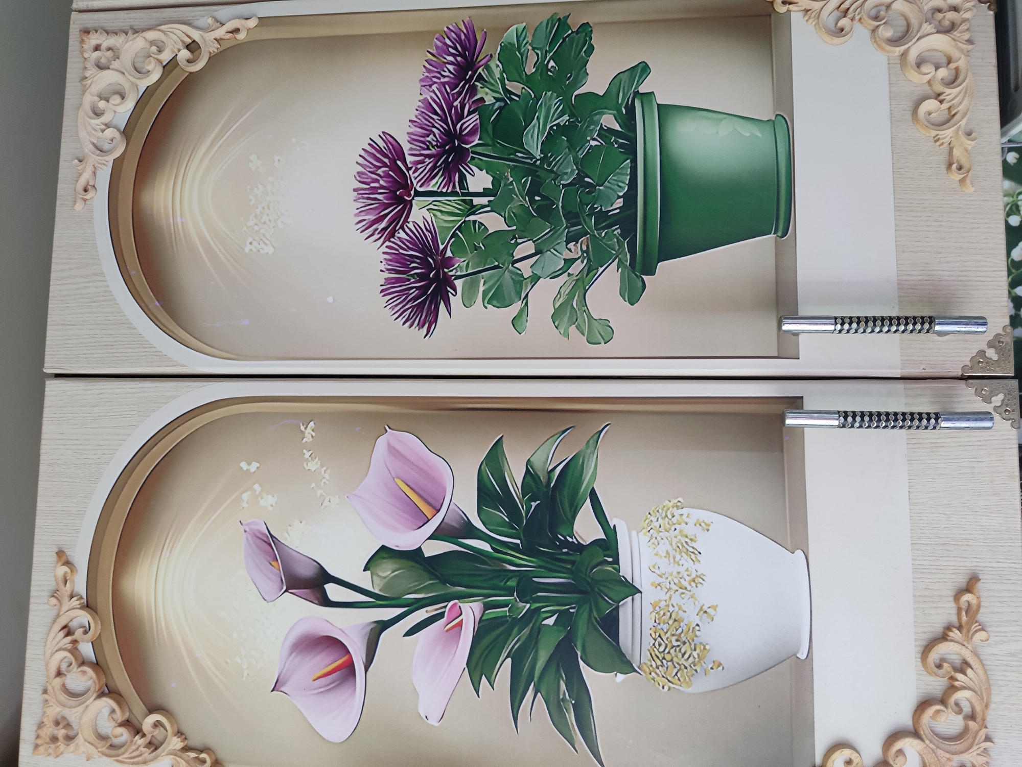 The image shows the interior of a cabinet with two decorative panels, each featuring a potted plant with a floral motif. The left panel displays a bouquet of pink calla lilies in a white vase adorned with yellow embellishments. The right panel showcases a cluster of purple asters in a green pot. Both panels are framed by arched borders with decorative wooden trim at the top corners. The cabinet has light-colored doors with silver handles. The overall aesthetic is ornate and traditional. The image is of an interior space, likely a home or a shop, and there is no visual information to connect it to the city of Noginsk, Russia.
