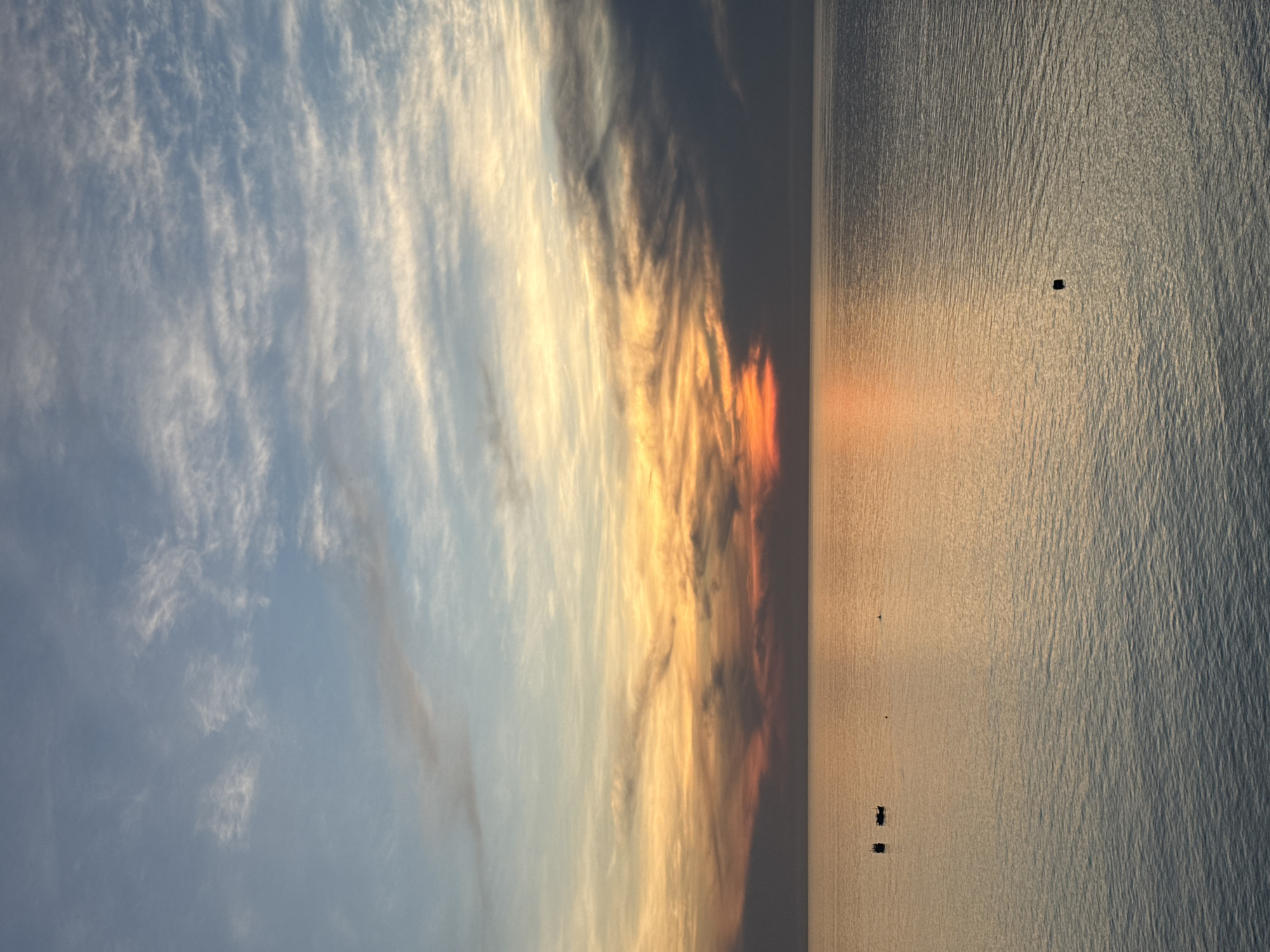 A serene sunset over the ocean at Lý Sơn Special Administrative Region, Vietnam. The sky is a canvas of muted blues, oranges, and yellows, with wispy clouds streaking across the horizon. The ocean, reflecting the warm hues of the sunset, is calm and vast, with subtle ripples disturbing its surface. Two small boats are visible in the distance, their dark silhouettes stark against the shimmering water. One boat appears to be closer to the shore, while the other is further out. In the foreground, a solitary black buoy floats on the water, a silent sentinel in the tranquil expanse. The overall scene evokes a sense of peace and solitude, a perfect moment captured as day transitions into night.