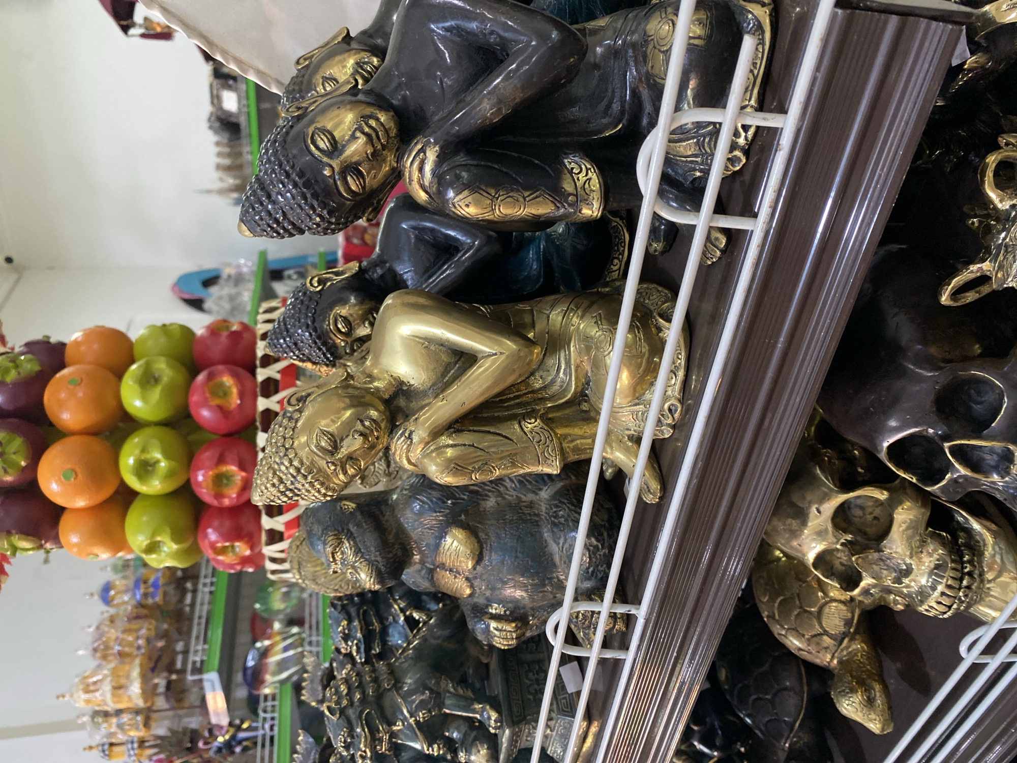 A horizontally-oriented close-up shot taken indoors shows rows of merchandise on shelves in a store. The location is Nusa Dua, Indonesia.
The items are on a white wire rack with a brown border. The top shelf has stacked, reclining Buddha statues. The next shelf contains skulls and turtles. To the left, behind the wire rack, is a display of produce, including oranges, green apples, and red apples. Other items in the background are small tourist items.
The Buddha statues are stacked. The top Buddha is black with gold accents, while the second Buddha is gold. The heads of the Buddhas have small tightly formed curls.
The skulls have large eye sockets and teeth. The turtle's shells are detailed with patterns.