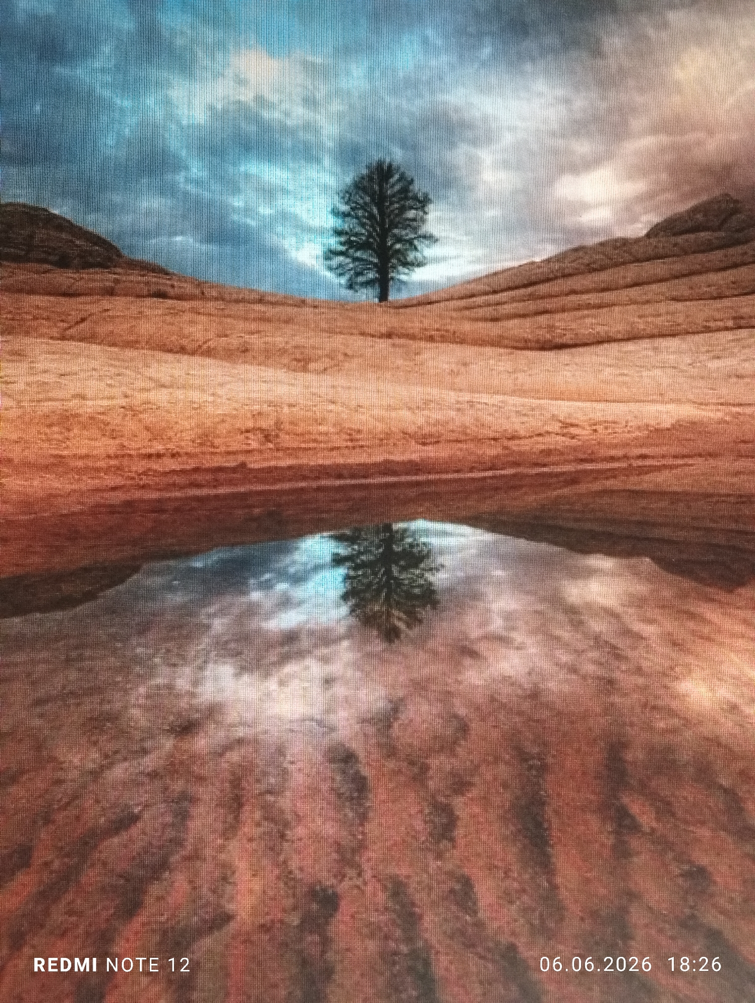 The scene depicts a tranquil landscape, set in the city of Burgas, Bulgaria, bisected horizontally by a still body of water that creates a clear reflection. In the upper half, a solitary, leafless tree stands centrally on the crest of a gently sloping, multi-layered rock formation. This formation exhibits prominent horizontal striations and rich reddish-brown hues, extending beneath an overcast sky composed of varying shades of blue, grey, and white clouds. The lower half of the image presents a near-perfect reflection of the tree, sky, and geological layers in the water. The immediate foreground, beneath the reflection, consists of similarly textured, reddish-brown ground. No people or animals are present. The entire image is overlaid with a visible grid-like pattern, characteristic of a photograph taken of a digital screen. White text overlays are present in the bottom corners: "REDMI NOTE 12" is in the bottom left, and "06.06.2026 18:26" is in the bottom right.