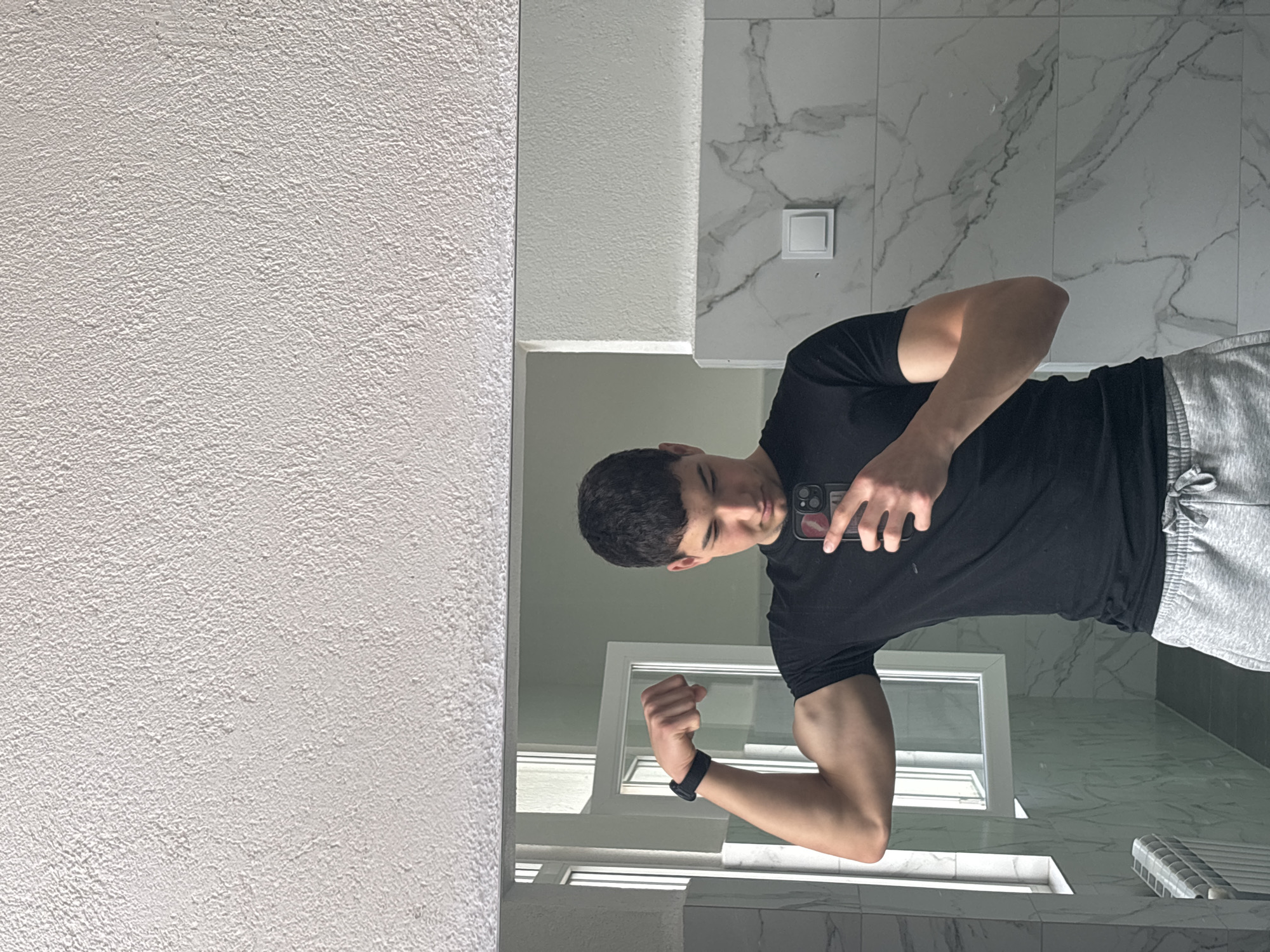 A young man takes a mirror selfie in what appears to be a modern bathroom in Zlatograd, Bulgaria. He is seen from the chest up, positioned slightly to the right of the frame, and is wearing a black t-shirt and gray sweatpants. His short, dark hair is styled, and he looks down towards his smartphone, which he holds in his right hand. A pink "lips" design is visible on the back of his phone case.
The man is flexing his left arm, showcasing his biceps, and has a black wristband on his left wrist. The reflection reveals a textured white wall on the far left, bordering the mirror. Behind the man, the bathroom features white tiled walls with subtle gray marble-like veining. A white square light switch is mounted on this tiled wall. Below the switch, a strip of light or reflective material is visible. Further down, a white-framed window reflects natural daylight into the room, and a white slatted object, possibly a radiator or vent, can be seen in the lower right corner of the reflection. The overall impression is a well-lit, contemporary interior space. The time of day appears to be daytime due to the natural light.