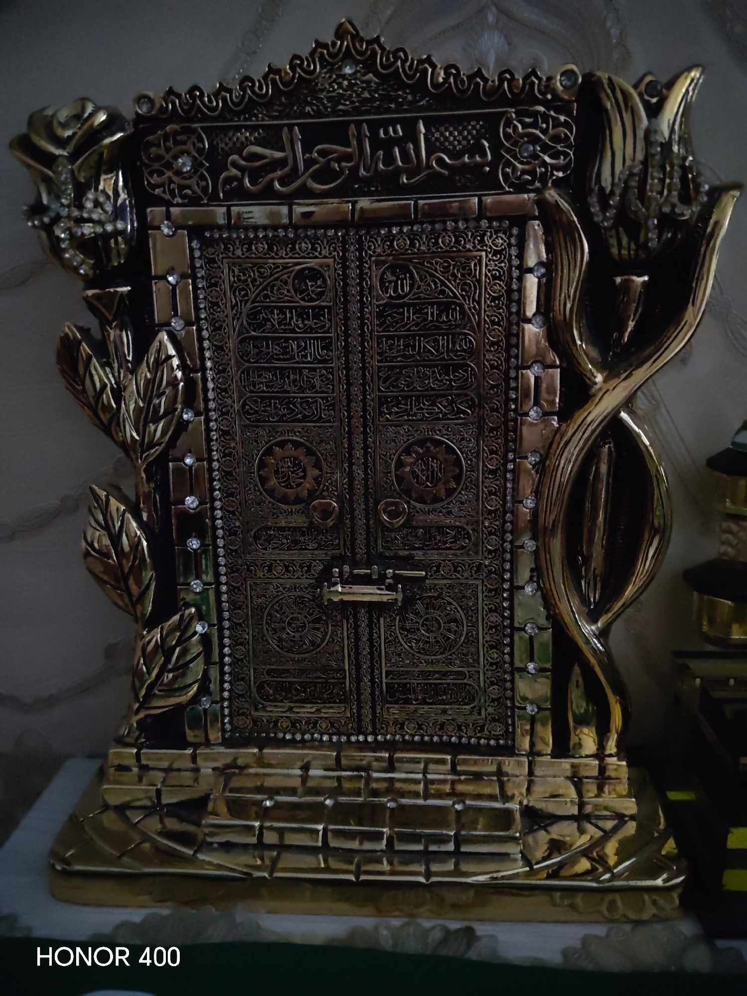 An ornate, gold-colored replica of the Kaaba door stands as the central subject, intricately decorated with Arabic calligraphy and embellished with numerous small, clear rhinestones. The replica is framed by decorative elements resembling large leaves on the left and a stylized, twisted metallic structure on the right. The base of the replica is designed to resemble stone steps. The overall setting appears to be indoors, with a textured wallpaper or fabric visible in the background. There are no people visible in the image. The bottom left corner of the image displays the text "HONOR 400". The provided crops focus on various details of the replica and its surroundings, including close-ups of the calligraphy, the rhinestones, and the decorative framing.