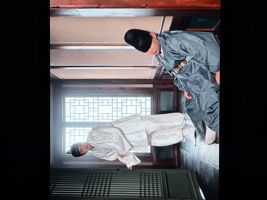 Two men wearing traditional Korean clothing are positioned within a room featuring a wooden floor and a window with a latticed design. One man, dressed in white, bows deeply. The other man, adorned in a gray garment and a black hat, bows slightly, with his gaze directed downward. The room's interior exudes a sense of traditional Korean architecture.
