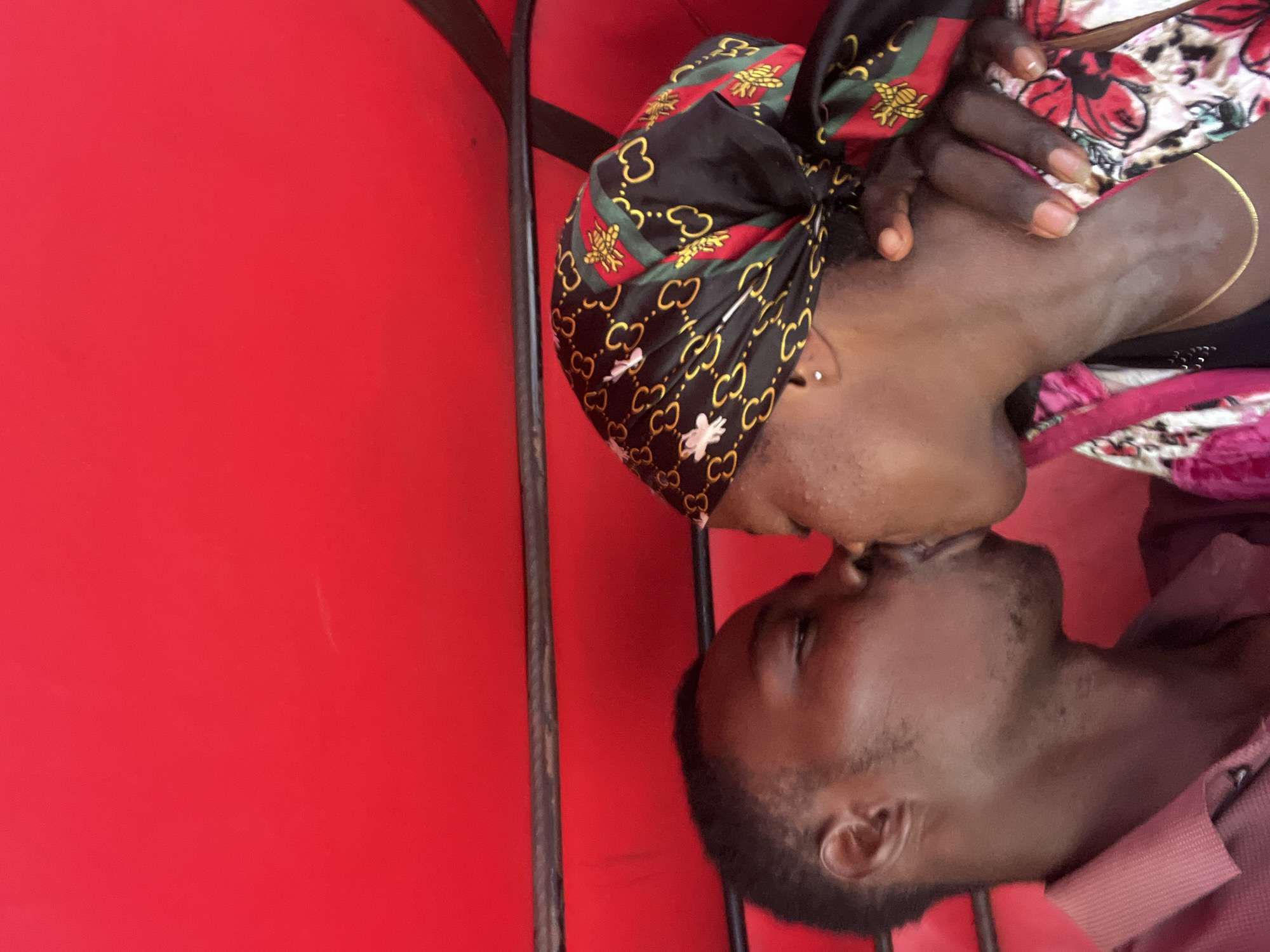 In Nkpor, Nigeria, a man and a woman share a kiss inside a vehicle. The woman is wearing a black headscarf patterned with gold interlocking G logos and red and green stripes, as well as a colorful jacket with floral prints. The man is wearing a red collared shirt. The interior of the vehicle is a vibrant red.