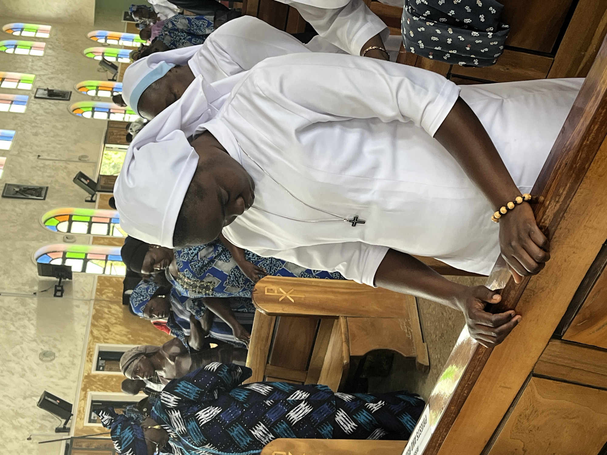 In the city of Vom, Nigeria, inside a church, a group of people are gathered in prayer. The primary focus is a young woman, a nun, wearing a white habit and veil. She is looking down, her hands clasped together on the wooden pew in front of her. A silver necklace with a cross pendant hangs around her neck, and a beaded bracelet adorns her wrist. To her right, another nun in a similar habit is partially visible. Other individuals, mostly women, are also present, some wearing colorful headscarves and patterned clothing, standing or sitting in the pews. The church interior features wooden pews, stained-glass windows with arched tops depicting various colors, and framed pictures on the walls. The overall atmosphere suggests a solemn religious service.