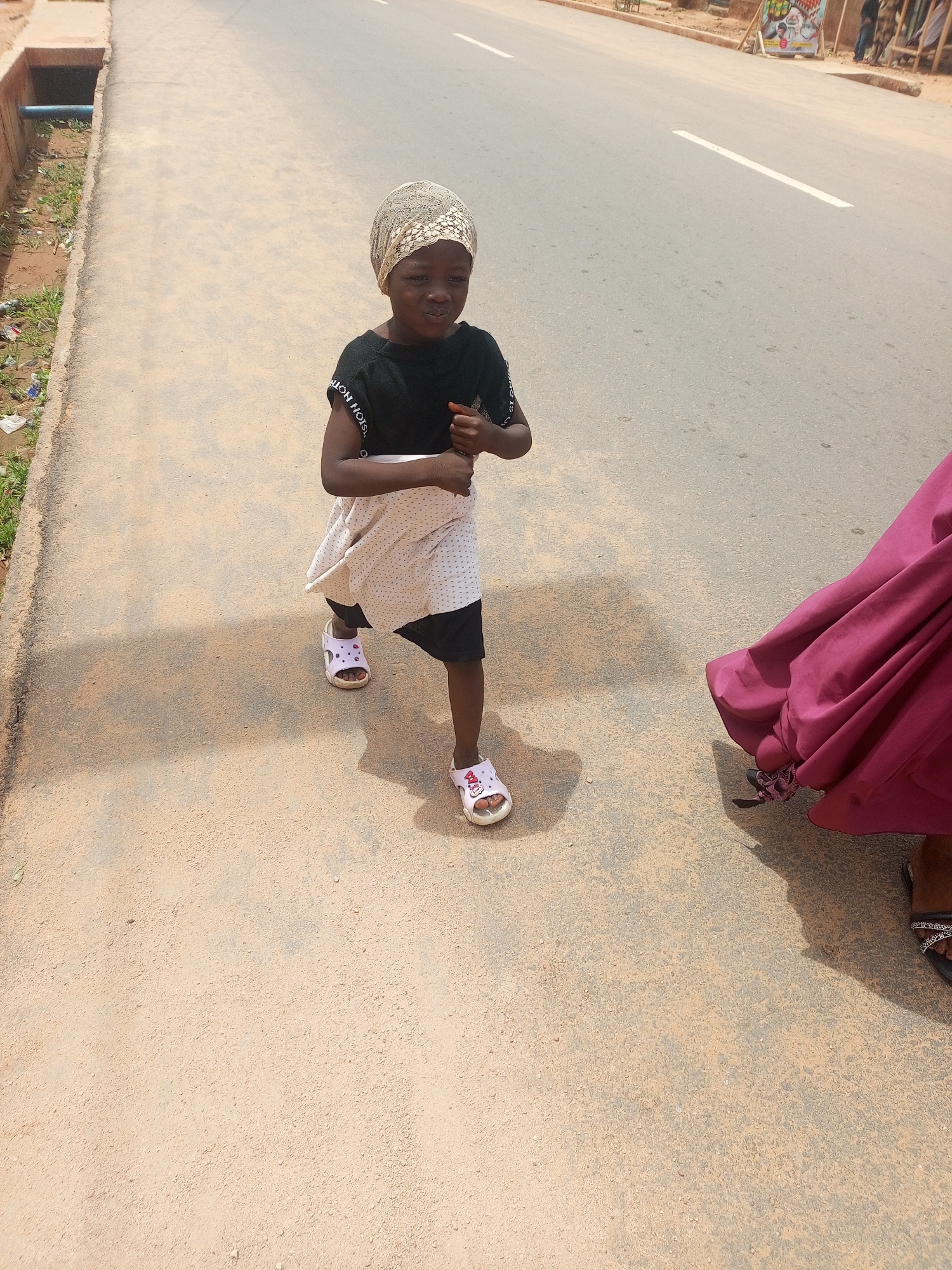 An outdoor scene captured from a slightly elevated perspective shows a young child walking on a paved surface, likely a sidewalk or road. The child, with dark skin, wears a light-colored, patterned head covering, a black short-sleeved top with text ("SION NOIS" - likely reversed "FASHION NOISE") on the sleeve, a light-colored, polka-dotted overlay skirt, and black bottoms underneath. White and pink open-toed sandals, possibly featuring a cartoon character design, are on their feet. The child's face is turned towards the camera with a slight pouting expression, and their arms are bent at the elbows, hands lightly clenched.
To the right of the child, the lower portion of an adult, wearing a flowing, dark magenta garment and dark sandals with embellished straps, is partially visible. A large shadow is cast on the ground from this individual.
The ground surface is a light-colored asphalt or concrete, showing wear and minor debris. On the left, a concrete curb separates the walking area from a lower drainage channel containing a blue pipe, sparse vegetation, and refuse. To the right of the child, a darker asphalt road is visible with a broken white line marking. In the distant background, structures, possibly buildings or market stalls, are present, along with a colorful sign on the right side of the road. The scene is brightly lit by natural sunlight.