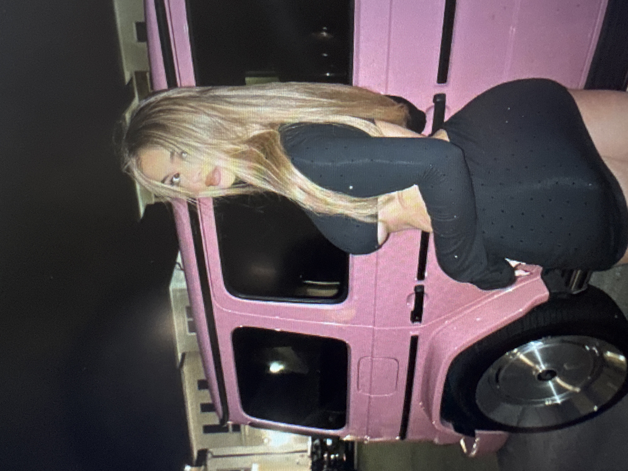The original image is rotated 90 degrees counter-clockwise. It depicts a woman posing alongside a vibrant pink vehicle, likely a Mercedes-Benz G-Wagen, in an outdoor nighttime setting. The woman has long, straight blonde hair and is wearing a form-fitting black garment, possibly a jumpsuit or dress, which features a pattern of small, shimmering perforations or embellishments. She is leaning against the passenger side of the SUV, with her body angled and her head turned towards the viewer. Her face, made up with visible eyeliner and lip color, holds a neutral to slightly alluring expression.
The bright pink G-Wagen has dark, tinted windows and black trim accents. A silver, reflective wheel with a smooth hubcap is visible at the bottom right of the frame. The background is dark and indistinct, indicating that the photo was taken at night, with artificial light sources illuminating the subjects. The overall scene conveys a sense of modern style and glamour. There is no visible text in the image.
Location context: Unknown, Unknown