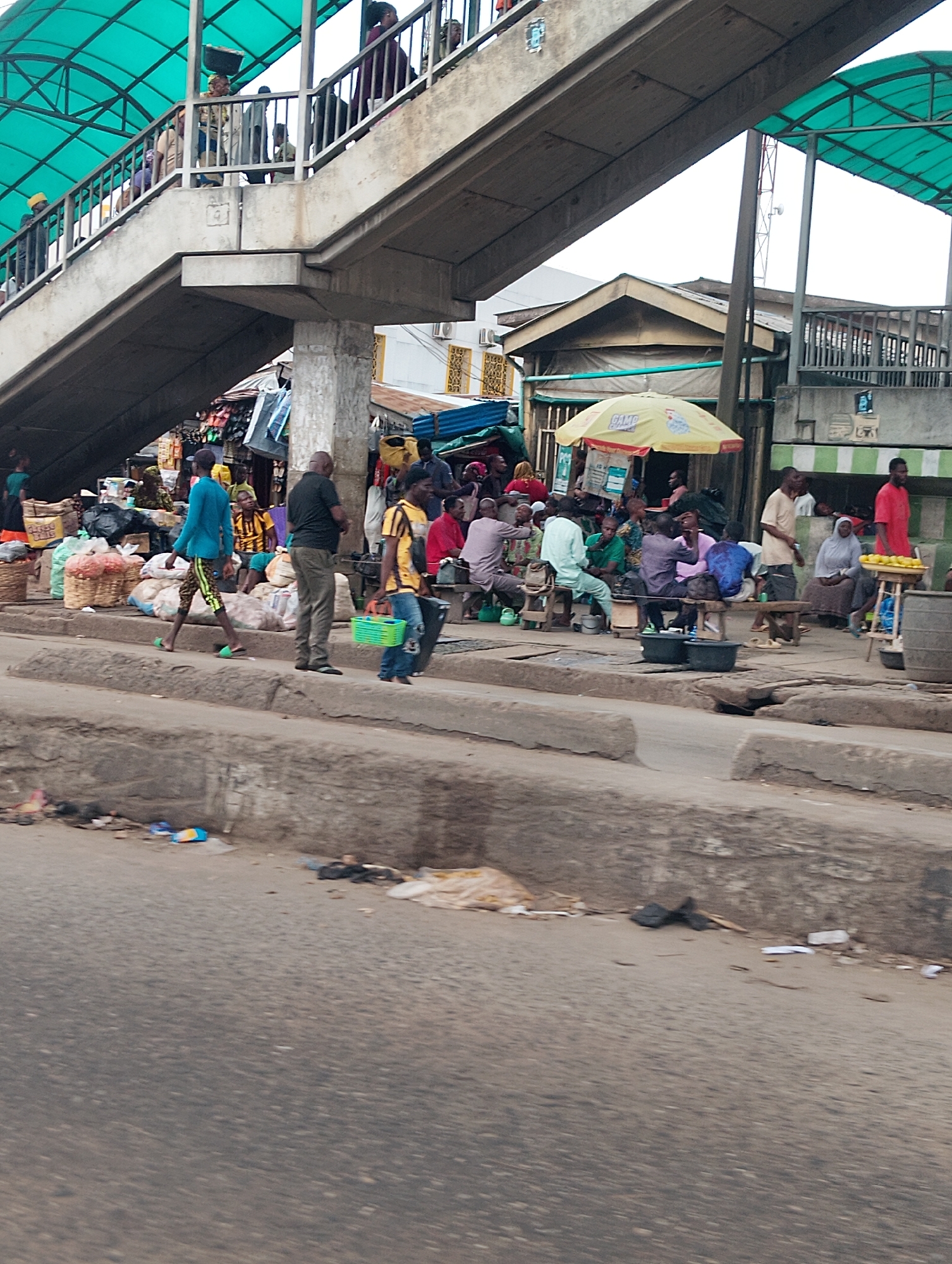 This media file depicts a bustling street scene in Shangisha, Nigeria, likely during daylight hours given the illumination. The foreground is dominated by a road with a raised concrete curb, on which several individuals are present. A man in a blue shirt and patterned shorts walks from left to right, his gaze directed forward. To his right, a man in a black shirt and grey pants stands facing away from the camera. Another man, wearing a yellow and black striped shirt, walks from right to left, carrying a green basket.
In the midground, a covered pedestrian walkway stretches across the frame. People can be seen walking on the walkway, some carrying items. Below the walkway is a market area. Stalls are set up, some with goods displayed, and several people are gathered, possibly shopping or socializing. A yellow umbrella provides shade over one of the stalls. Various objects are visible, including baskets of produce, bags, and what appear to be food items on a table. The environment suggests an active commercial hub.
The background reveals more of the market area and buildings. The overall atmosphere appears busy and typical of a Nigerian marketplace, with a mix of shoppers, vendors, and pedestrians navigating the space. The weather appears to be fair, with no visible signs of rain. The scene is vibrant with human activity, highlighting the everyday life in this location. There is no visible text in the provided image.