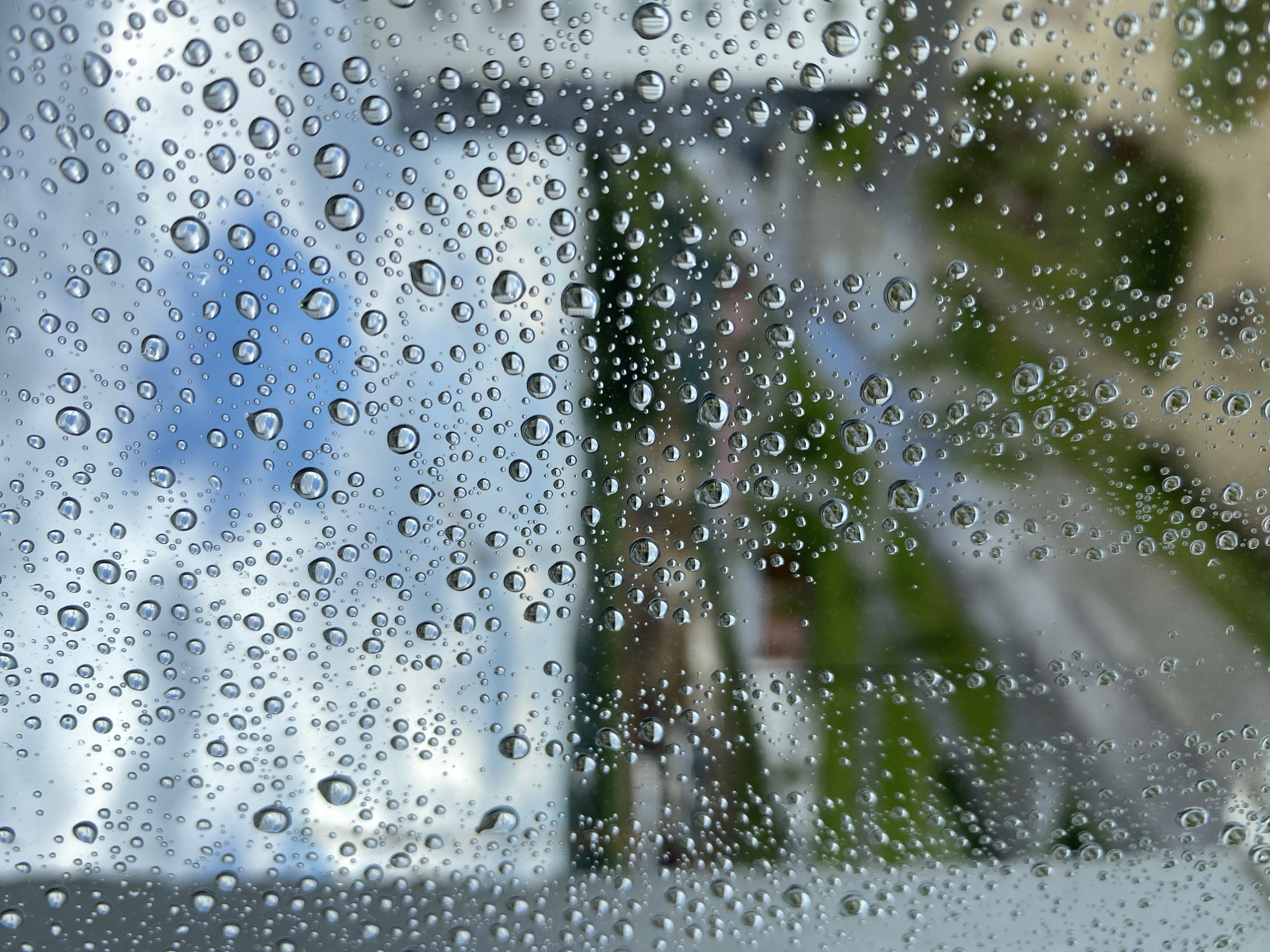 The image depicts a view through a window covered in numerous raindrops, with the city of Yekaterinburg, Russia, blurred in the background. The foreground is dominated by water droplets of varying sizes, which distort the scene beyond. The background shows a mixture of muted green foliage and urban structures, including buildings with red and muted-colored roofs, and what appears to be a paved area or road. The sky above is partly cloudy, with patches of blue visible through the overcast conditions. There are no people or active events visible in the image; it is a static, atmospheric shot of a rainy day.