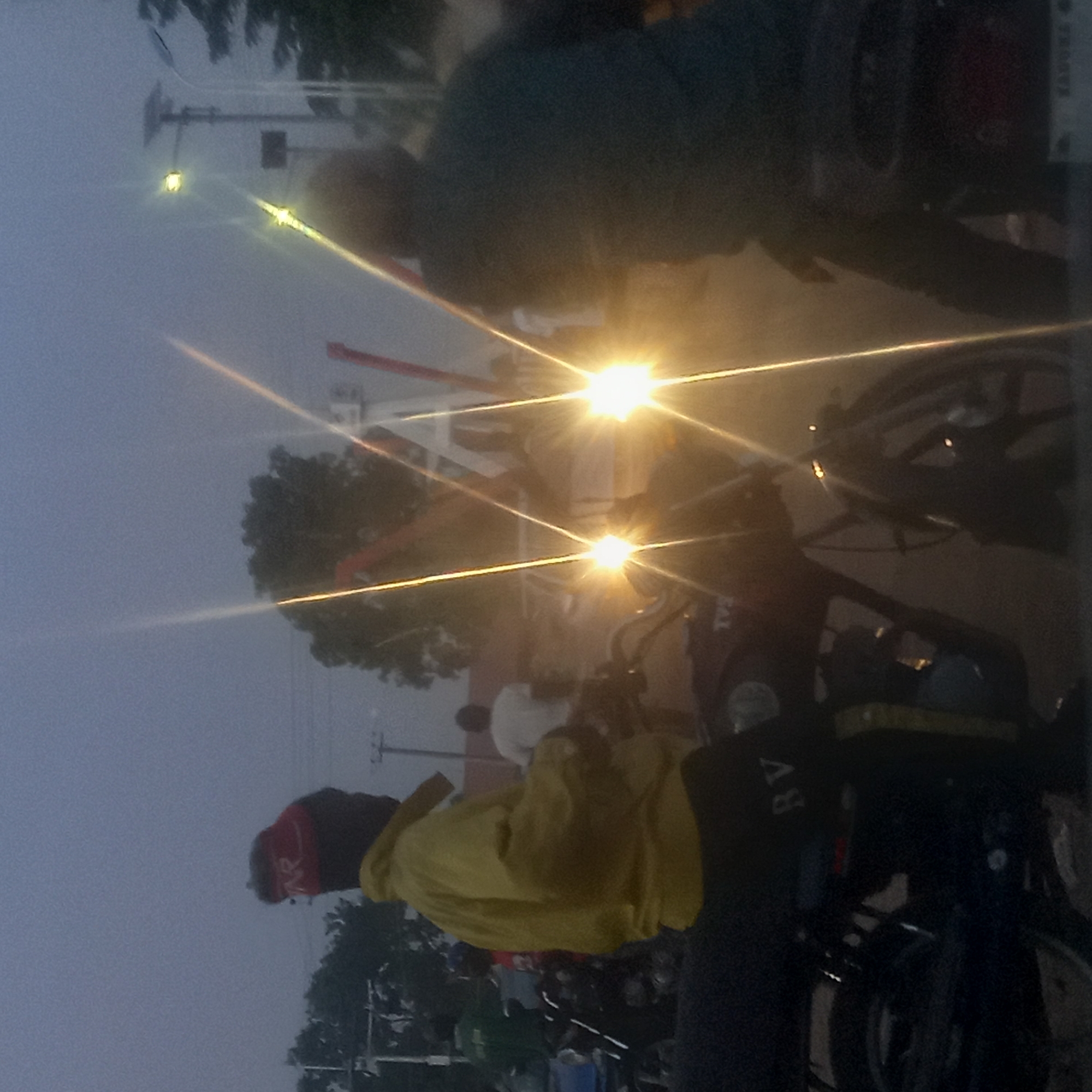 The image depicts a street scene in Kinkinau, Nigeria, at dusk or dawn, possibly during heavy fog. Several people on motorcycles are present, with headlights illuminating the scene. One individual wears a yellow jacket and a red head covering. Some infrastructure such as street lights and trees are visible in the background. The overall environment appears hazy.