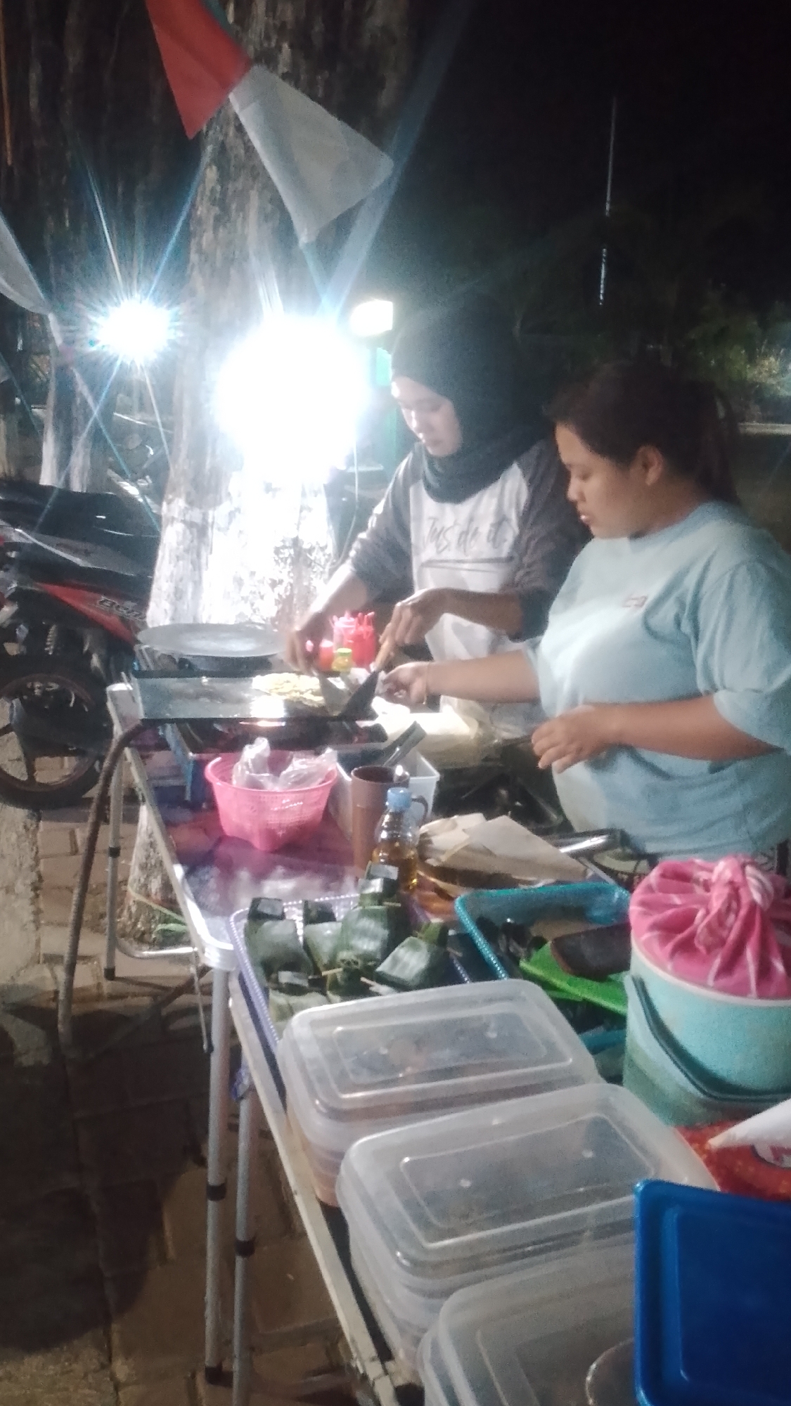 In the city of Karanglangu, Indonesia, at night, two women are preparing food at a street stall. One woman, wearing a black hijab and a grey and white shirt, is cooking on a griddle with a spatula. The other woman, wearing a light blue t-shirt, is standing beside her, holding a spatula and looking at the food. The stall is set up on a table covered with a patterned cloth. On the table, there are various food items, including wrapped parcels in banana leaves, plastic containers filled with food, and a pink basket. A motorcycle is parked to the left of the stall. Bright lights are illuminating the scene, creating glare. Flags are visible in the background. The ground appears to be paved with bricks.