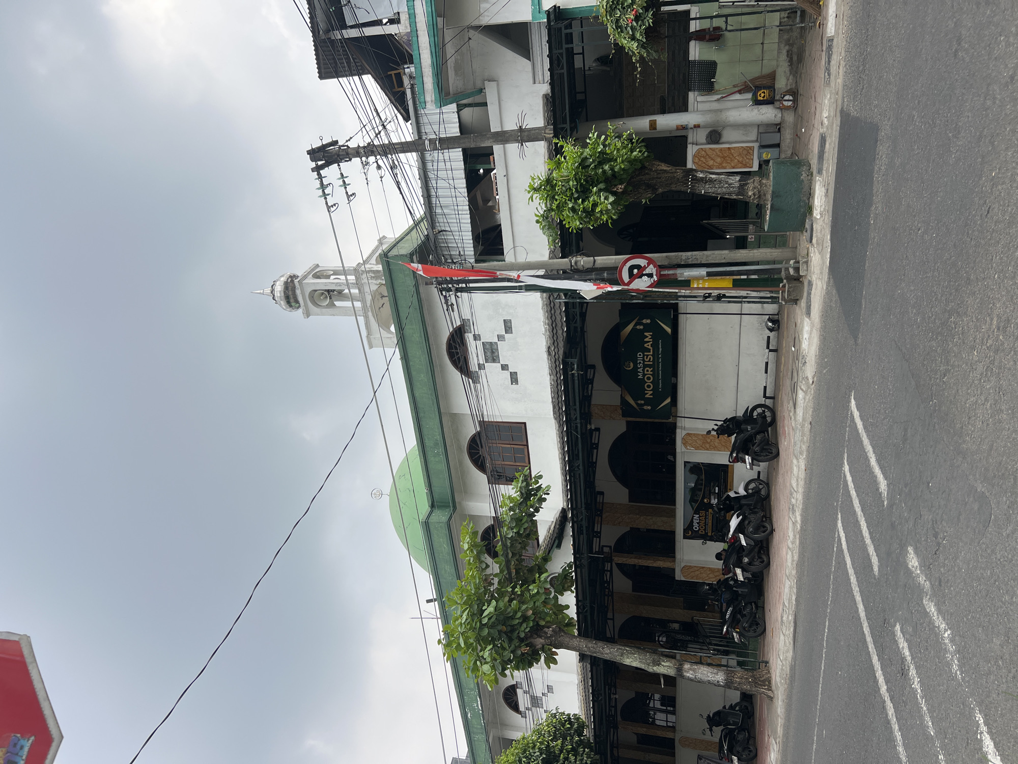 A white mosque with a green dome, named "MASJID NOOR ISLAM", is situated in Yogyakarta, Indonesia. The mosque has a tall, white minaret with a silver-capped dome, and a row of arched windows on its upper floor. In front of the mosque, a covered walkway with arched entrances is supported by pillars. Several motorcycles are parked along the sidewalk in front of the mosque. A street with crosswalk markings runs in front of the building. A red and white Indonesian flag is displayed on a pole next to a sign with a "no parking" symbol. The sky is overcast.