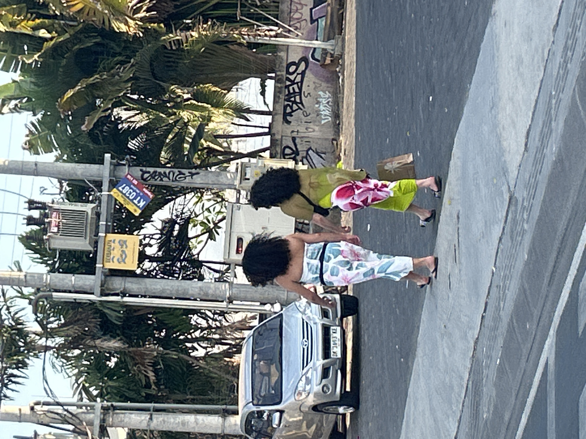 Two women walk on a sidewalk in North Kuta, Indonesia, with a silver car parked nearby. The woman on the left wears a strapless top and patterned pants, while the woman on the right is dressed in a green tunic over a patterned skirt and carries a shopping bag. Palm trees and electrical poles line the street, and graffiti adorns a concrete wall in the background. The scene is illuminated by bright sunlight, casting shadows on the ground.