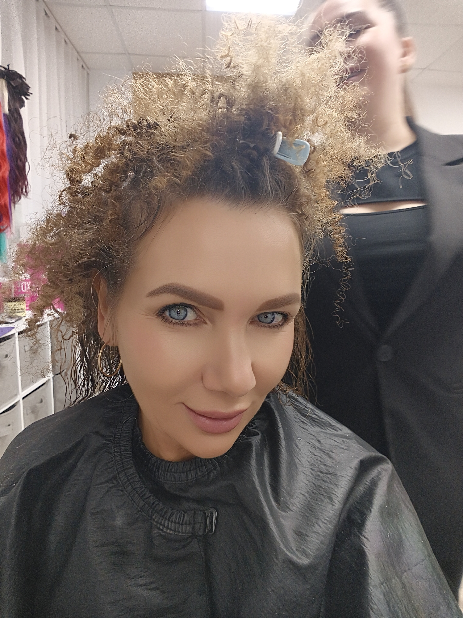 A woman with striking blue eyes and light brown, curly hair is the central focus of the image. Her hair appears wet and is styled with some sections clipped up on top with a light blue clip. She is wearing a black salon cape over her shoulders and has a subtle smile, looking directly at the camera. A single gold hoop earring is visible in her left ear.
In the background, a second person, out of focus, is partially visible behind the main subject's head, wearing a black top or jacket. This person's profile is obscured, but they appear to be standing or working behind the primary subject. To the far left, several colorful hair extensions or products hang from a rack. White, multi-drawer storage units are also visible in the background, further suggesting an indoor salon or beauty studio setting. The ceiling with overhead lighting is partially visible at the top. The overall scene depicts someone in the process of getting their hair styled or treated. No discernible text is visible.