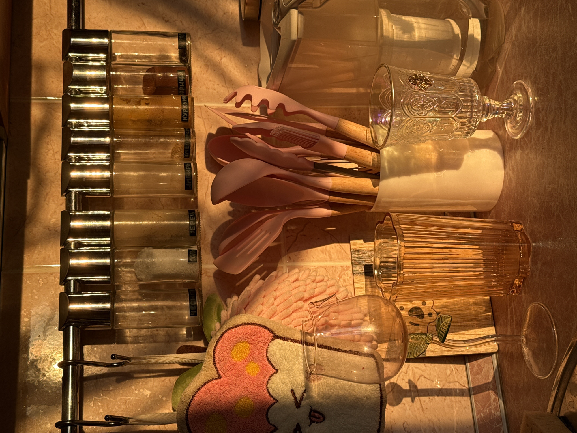 The image shows a close-up view of a kitchen counter bathed in warm, golden sunlight, creating long shadows. In the background, a rack holds several glass spice jars with metallic lids, some containing visible spices. To the left, a pink towel with a cartoonish cupcake design is hanging. In the foreground, a collection of kitchen utensils, primarily pink silicone with wooden handles, are held in a white ceramic container. In front of the utensils, a tall, ribbed amber glass stands next to a delicate, stemmed glass with a floral design. A clear, ornate glass goblet with a pedestal base is positioned to the right. The overall impression is of a cozy, domestic kitchen setting.
