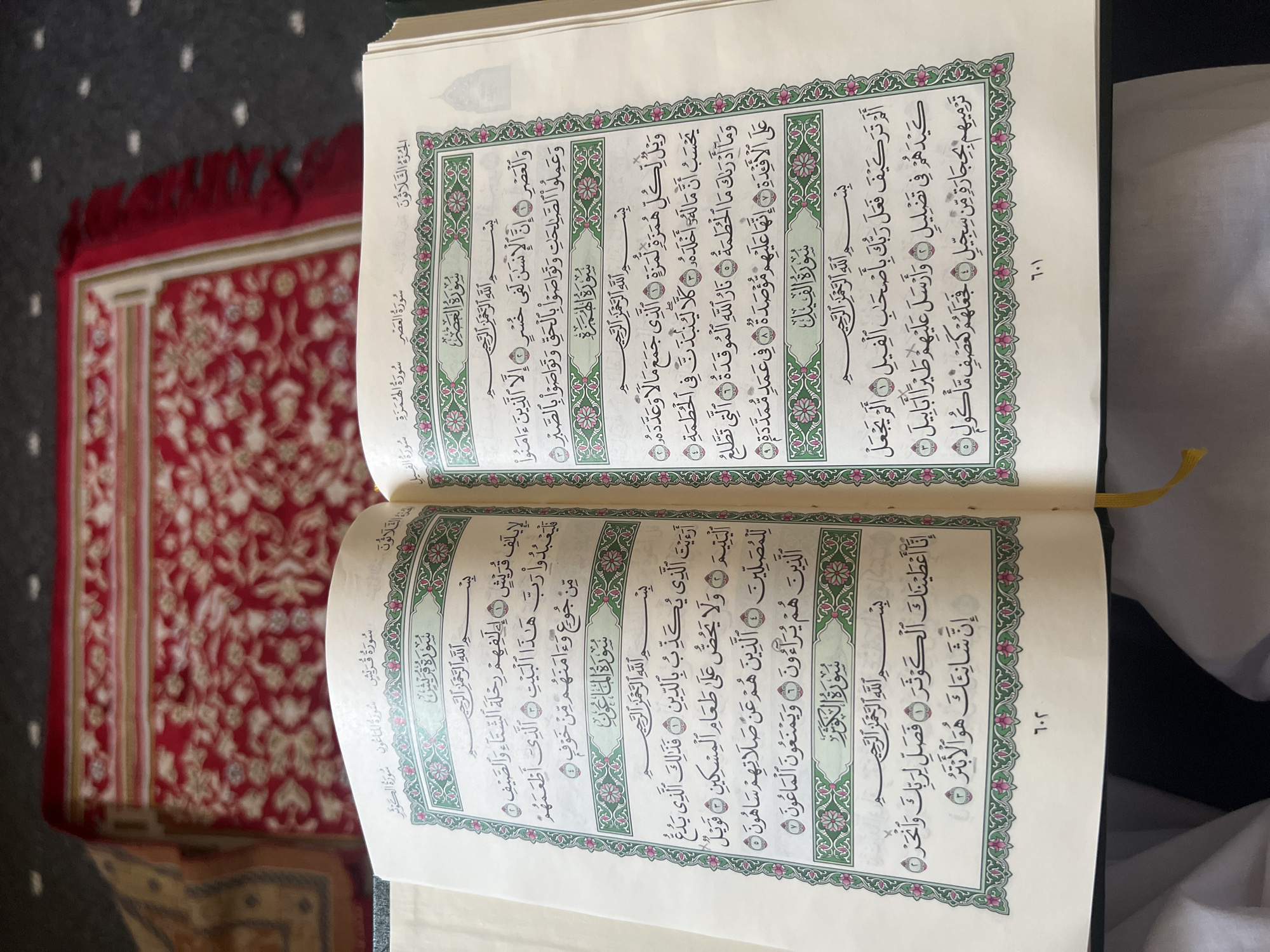 The image shows an open Holy Quran with Arabic text displayed on both pages, placed on a patterned red carpet. The carpet has a traditional Islamic design with intricate floral motifs and a fringed edge visible on the right side. The Quran is open to the section containing Surah Al-Qari'ah, Surah Al-Adiyat, Surah Az-Zalzalah, Surah Al-Inshirah, Surah At-Tin, Surah Al-Alaq, Surah Al-Qadr, Surah Al-Bayyinah, Surah Az-Zilzal, Surah Al-Adiyat, Surah Al-Qari'ah, Surah At-Takathur, Surah Al-Asr, Surah Al-Humazah, and Surah Al-Feel. The page numbers are visible at the bottom of each page, with 701 on the left and 702 on the right. The lighting suggests an indoor environment, possibly a mosque or a home. There are no people or specific landmarks that would indicate a precise location within Makkah Al Mukarramah, Saudi Arabia.